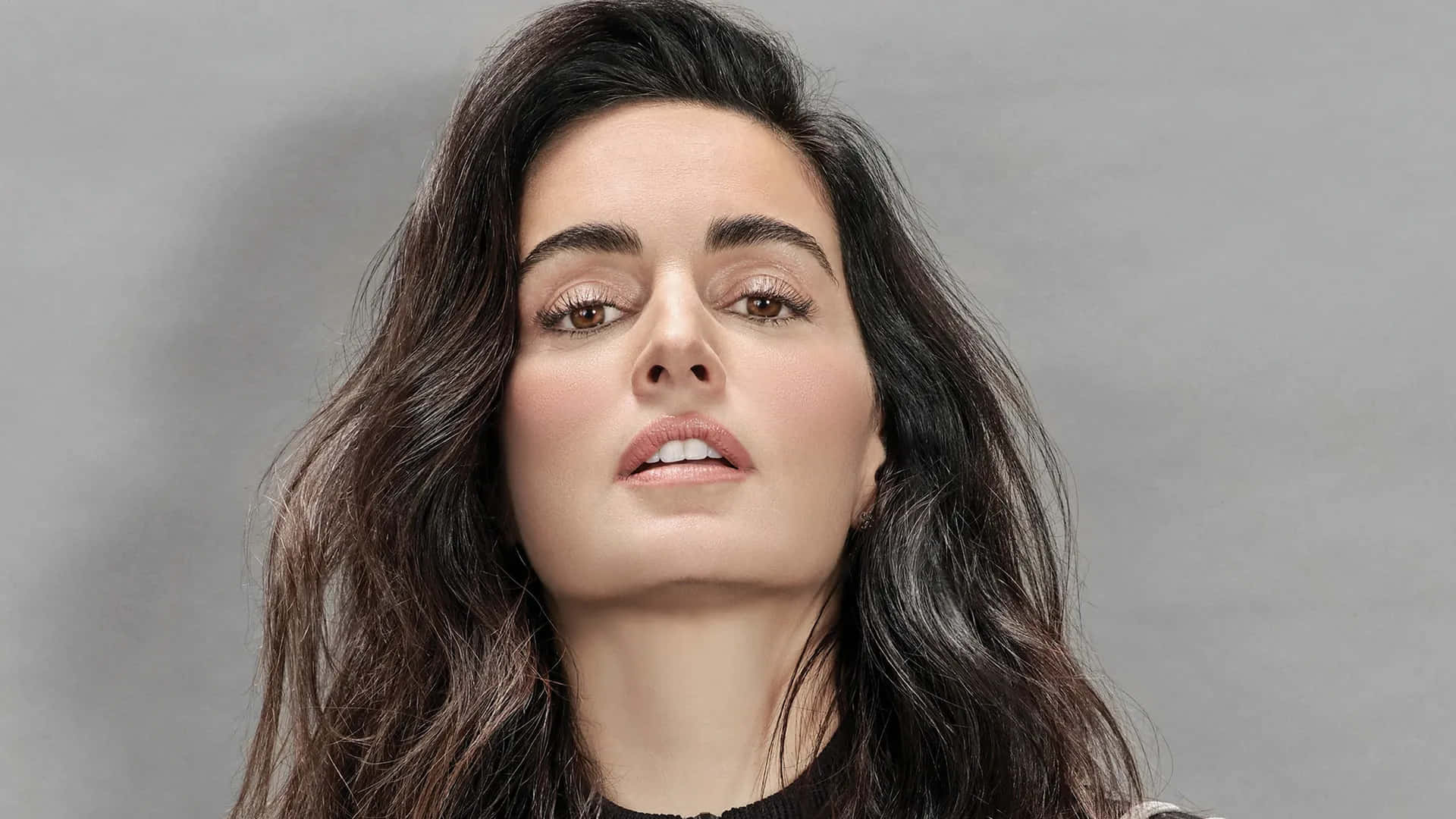 Ana De La Reguera 桌布 壁紙 — 一位擁有深色波浪長髮的女性在柔和的灰色工作室背景前向上凝視。