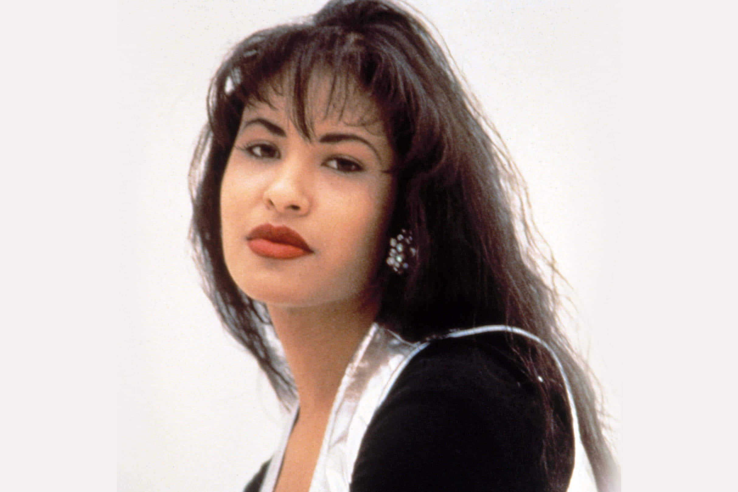 Selena Quintanilla iPhone 桌布 壁紙 — 一位擁有深色波浪頭髮和大膽紅唇的女性，在純白背景前自信地凝視。