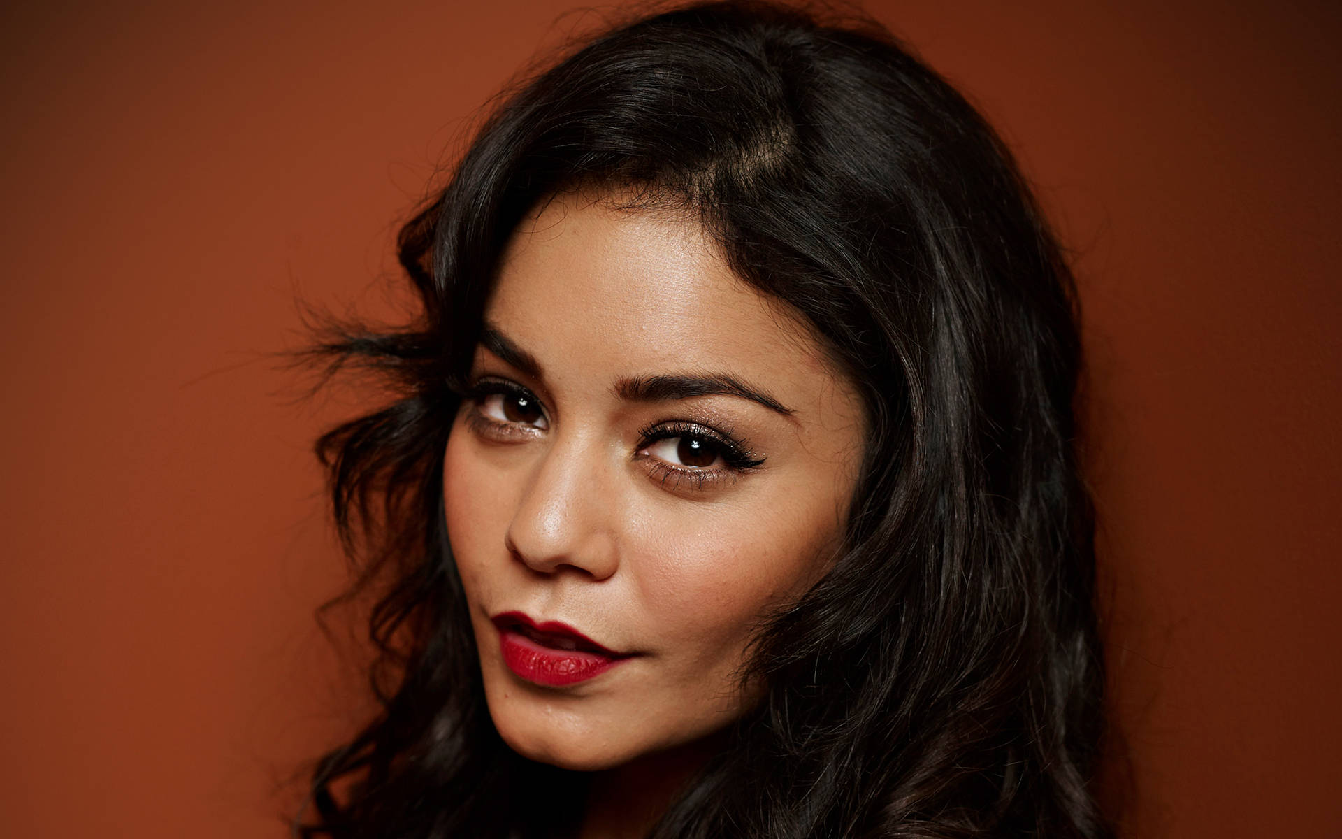 Vanessa Hudgens 桌布 壁紙 — 一位擁有深色波浪頭髮和大膽紅唇的女性，在溫暖的赤陶背景前直視前方。