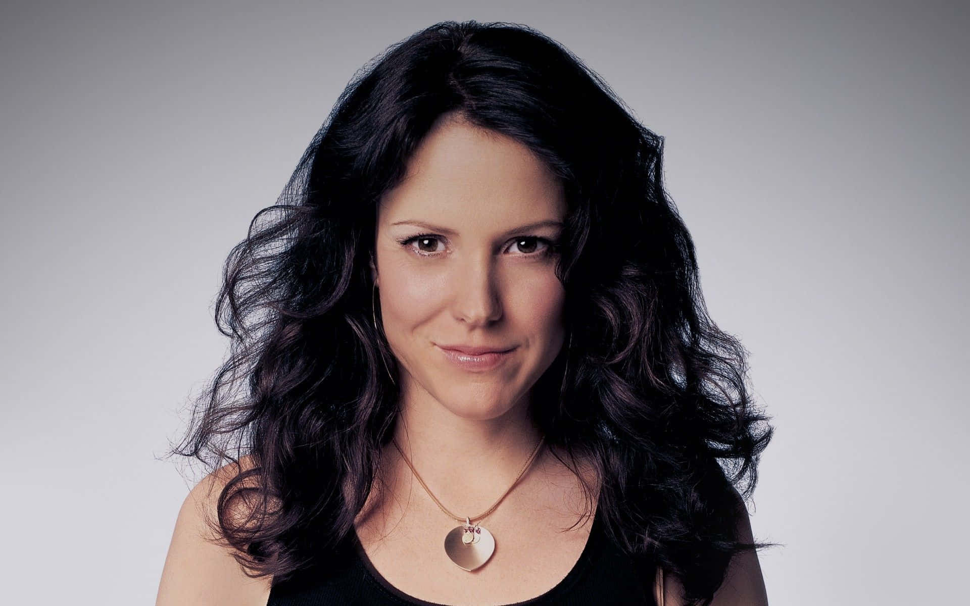 mary louise parker 桌布 壁紙 — 一位擁有深色波浪頭髮和心形吊墜的女性，在純灰色背景前溫柔微笑。