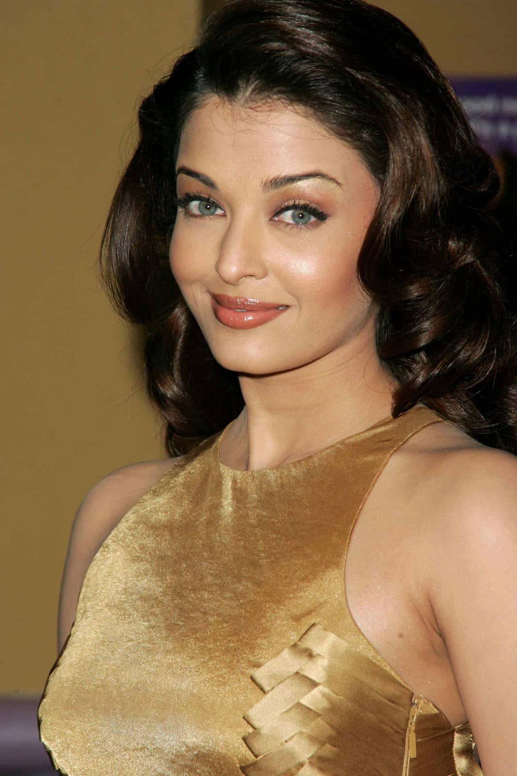 Aishwarya Rai 桌布 壁紙 — 一位擁有深色波浪髮和綠色眼睛的女性，穿著閃閃發光的金色無袖連衣裙，溫暖地微笑。