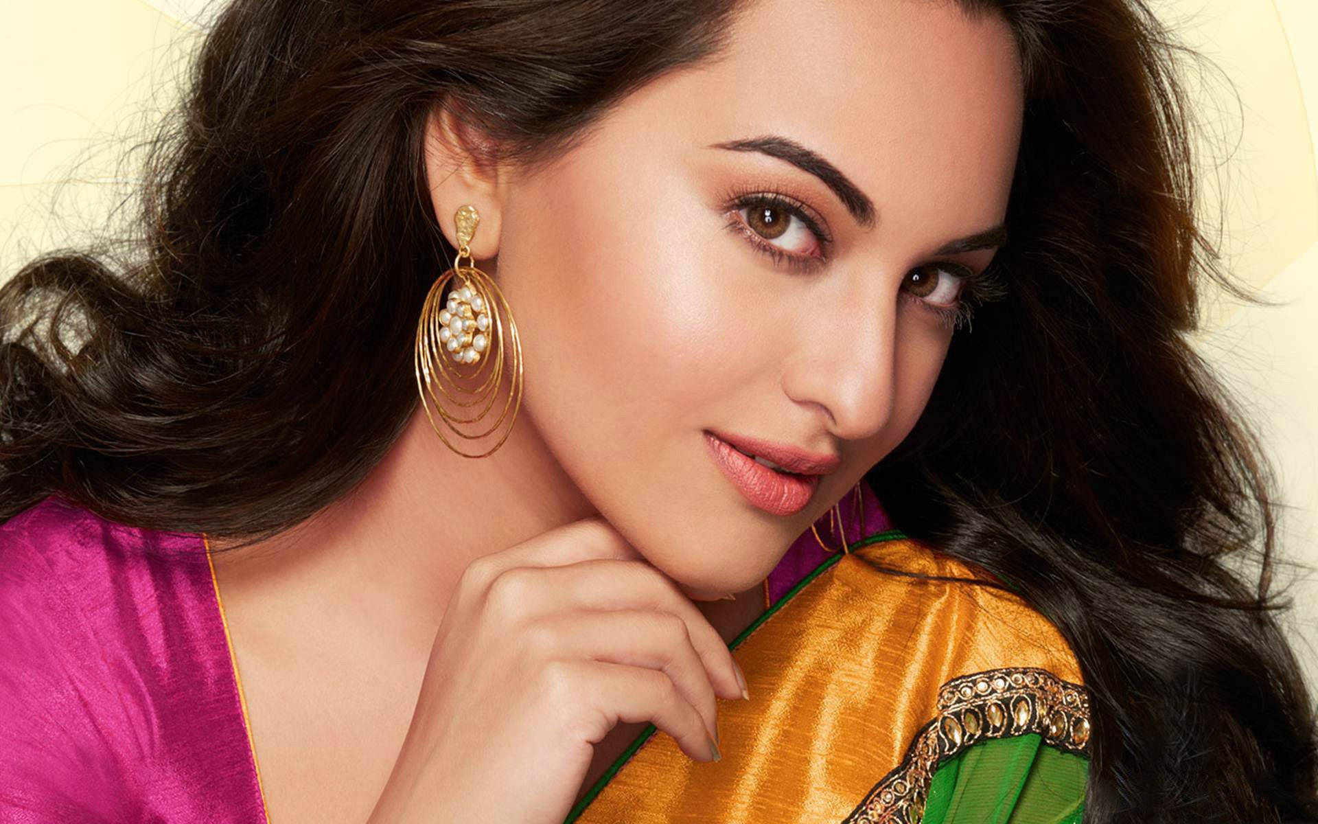 Sonakshi Sinha 桌布 壁紙 — 一位擁有深色波浪髮和金色耳環的女性，穿著鮮豔的粉紅色和金色紗麗，溫柔地凝視著。