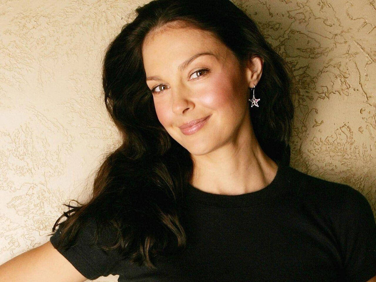 Ashley Judd 桌布 壁紙 — 一位擁有深色波浪髮型和星星耳環的女性，在紋理米色牆壁前溫柔微笑。
