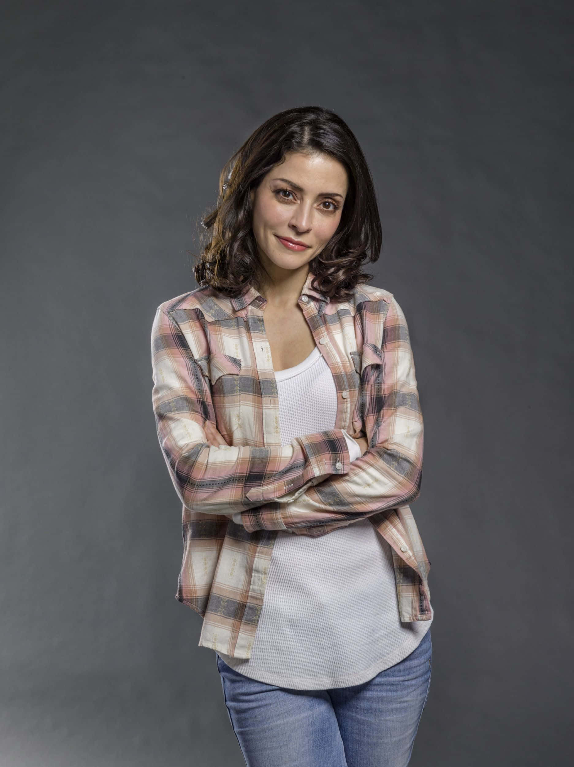 Emmanuelle Vaugier 桌布 壁紙 — 一位擁有深色波浪髮的女性自信地交叉雙臂站立，穿著格紋襯衫內搭白色上衣和牛仔褲，背景是灰色攝影棚。