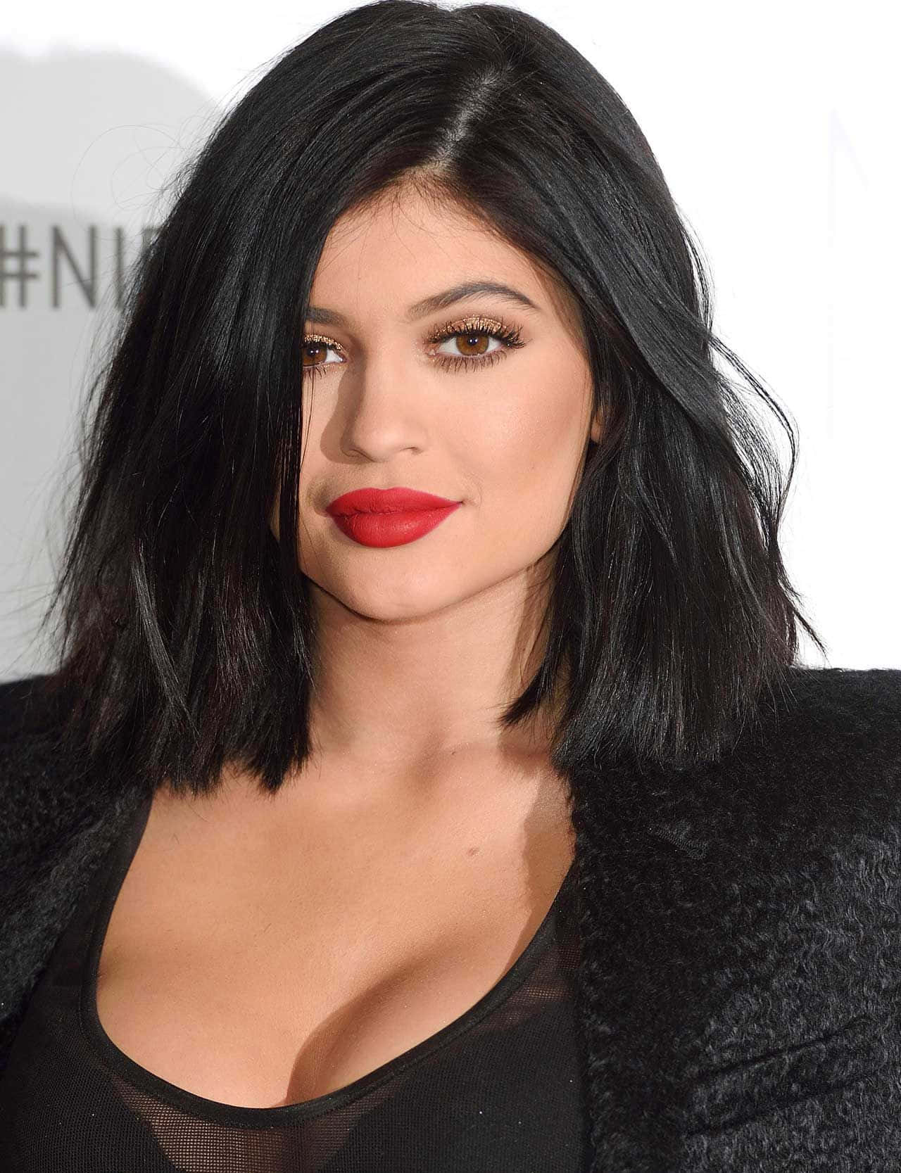 凱莉·珍娜 桌布 壁紙 — 一位擁有深色波浪鮑伯頭、大膽紅唇和煙燻妝眼睛的女性穿著黑色紋理外套的特寫肖像。
