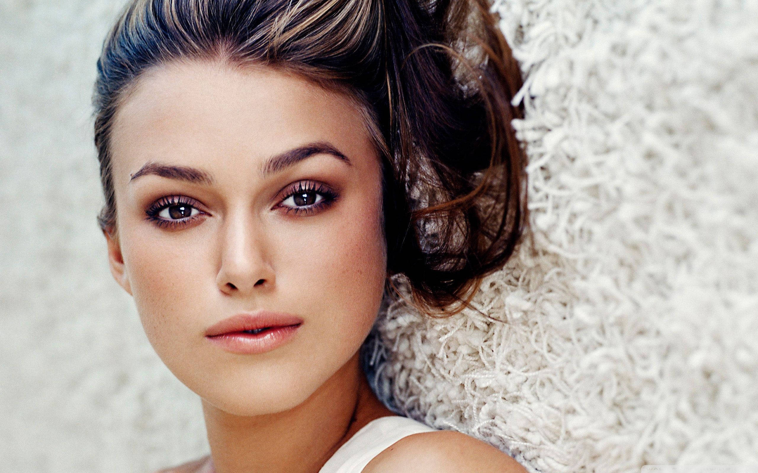 Keira Knightley 桌布 壁紙 — 一位擁有深色眼睛和造型頭髮的女性，依靠在蓬鬆白色紋理背景上的特寫肖像。
