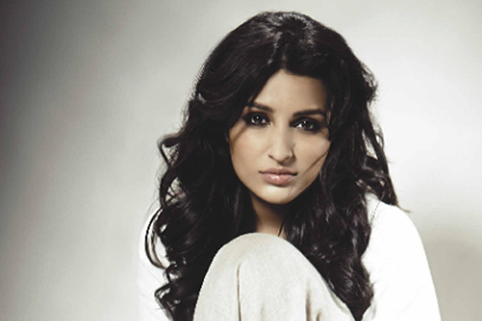 Parineeti 桌布 壁紙 — 一位擁有深色長波浪髮的女性在柔和的灰色工作室背景下直視鏡頭。
