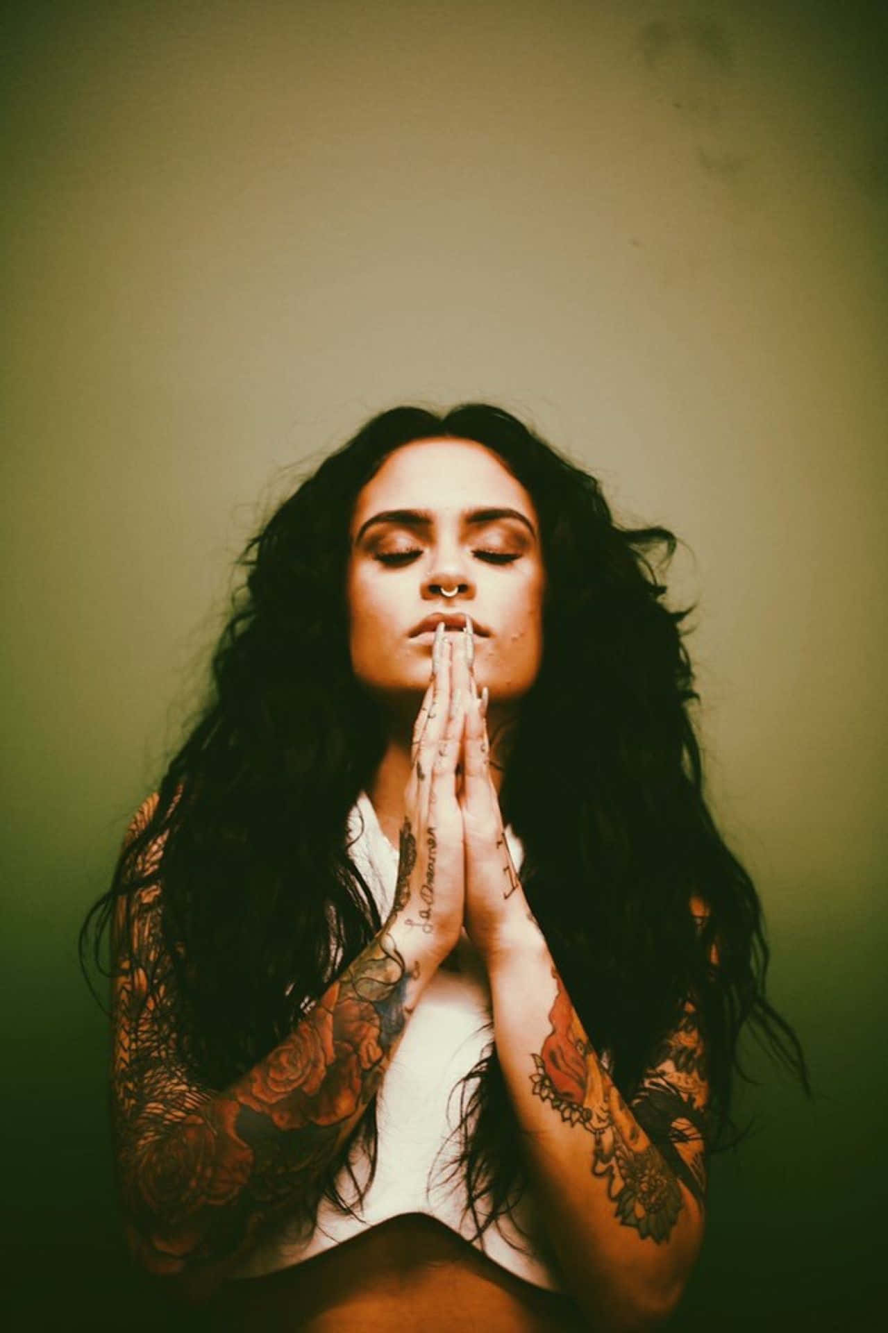kehlani 桌布 壁紙 — 一位擁有深色長髮和花卉刺青的女性，雙手合十靠近嘴唇，對著柔和的綠色牆壁祈禱。