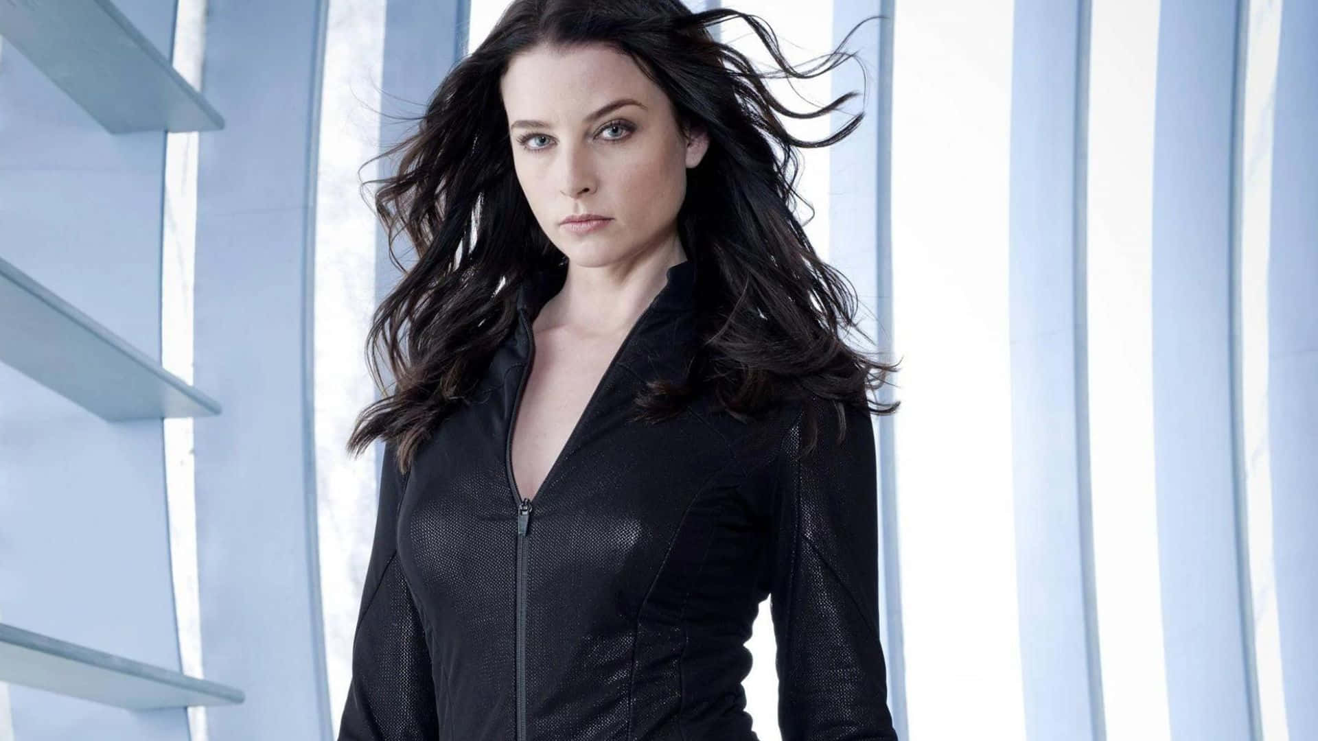 rachel nichols 桌布 壁紙 — 一位擁有深色飄逸長髮和銳利眼神的女性，站在現代藍色燈光的室內，身穿黑色拉鍊外套。
