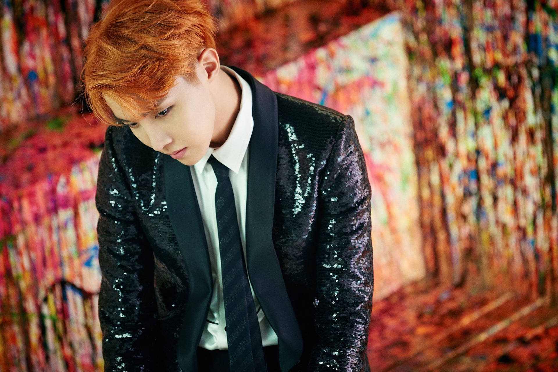jhope 桌布 壁紙 — 一位擁有火橙色頭髮的年輕男子，在充滿活力的抽象彩繪背景下，穿著閃亮的黑色西裝外套。