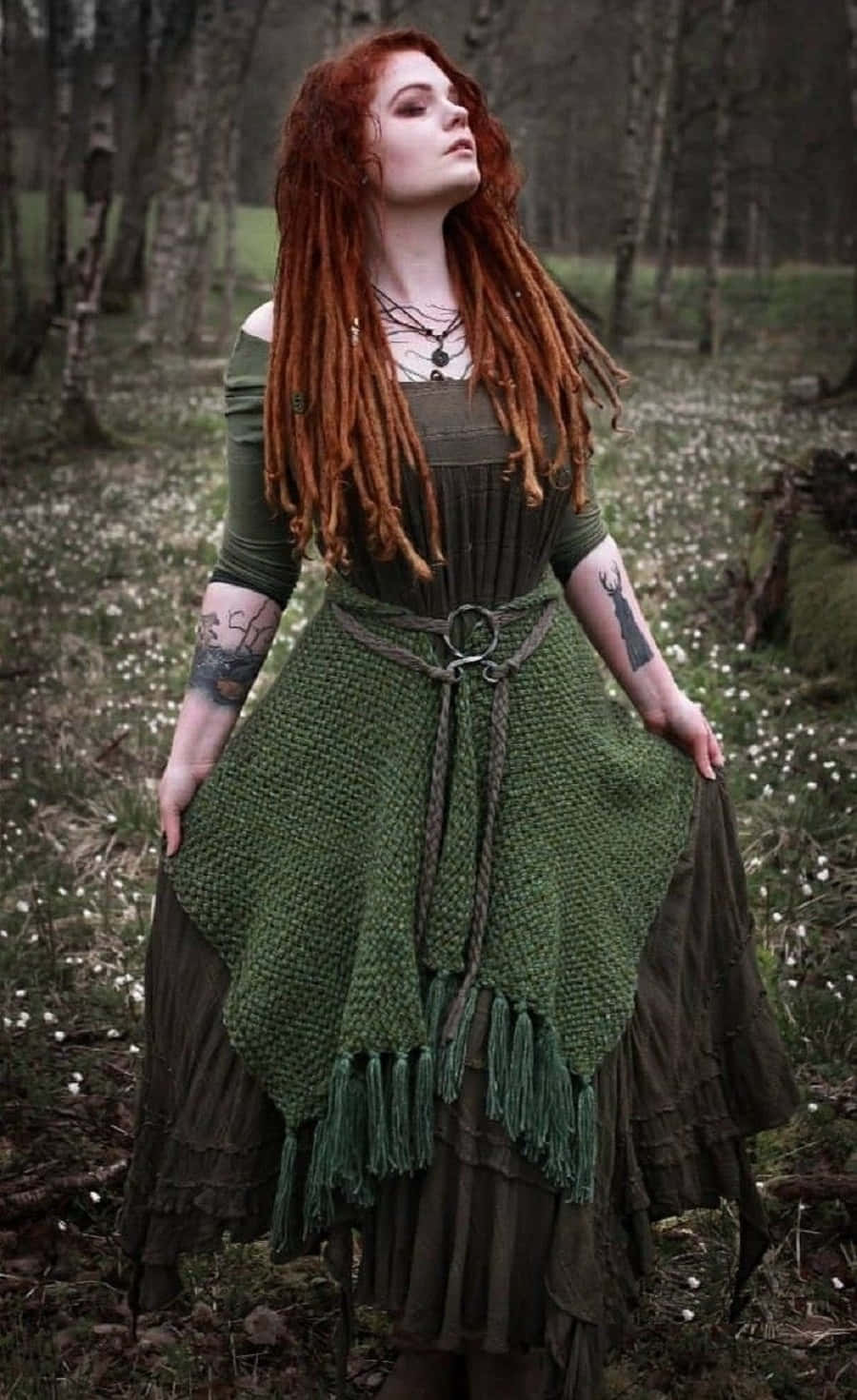 Witch Costumes 桌布 壁紙 — 一位擁有火紅辮子的女性站在迷霧森林中，身穿帶流蘇的土綠色分層連衣裙，手臂上可見刺青。