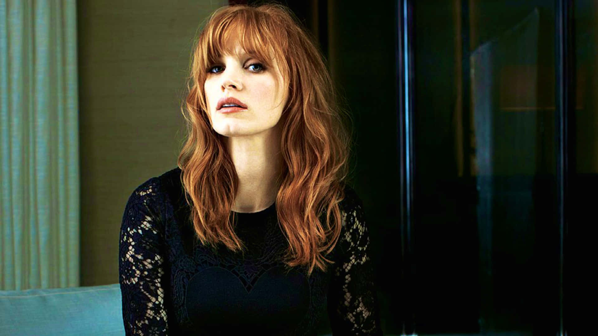 jessica chastain 桌布 壁紙 — 一位擁有火紅頭髮和強烈藍眼睛的女性在一個有窗簾和玻璃門的昏暗房間裡向上凝視。