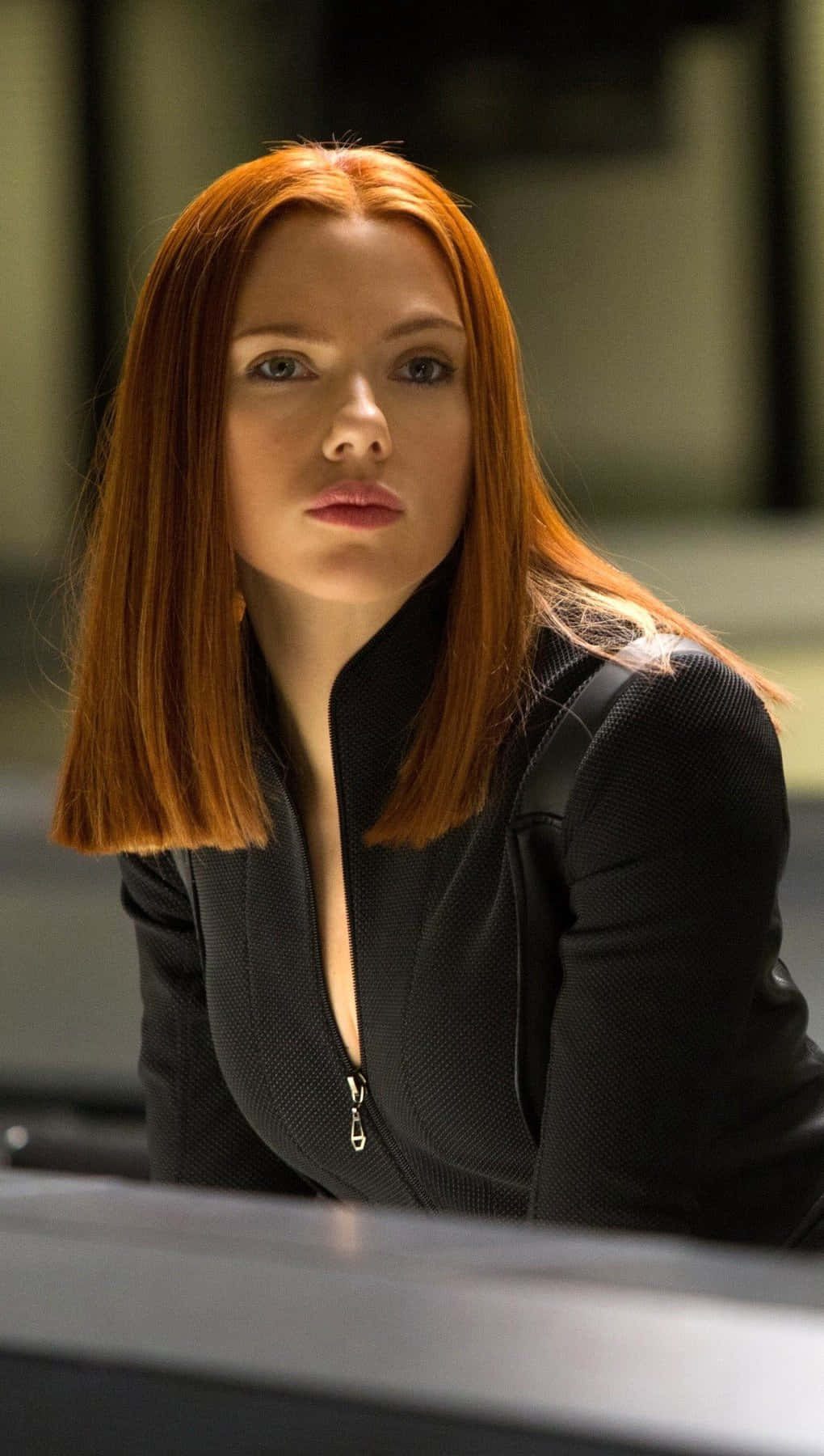 Natasha Romanoff 桌布 壁紙 — 一位擁有火紅頭髮和銳利眼神的女性，在燈光昏暗的現代室內穿著一件時尚的黑色戰術外套。