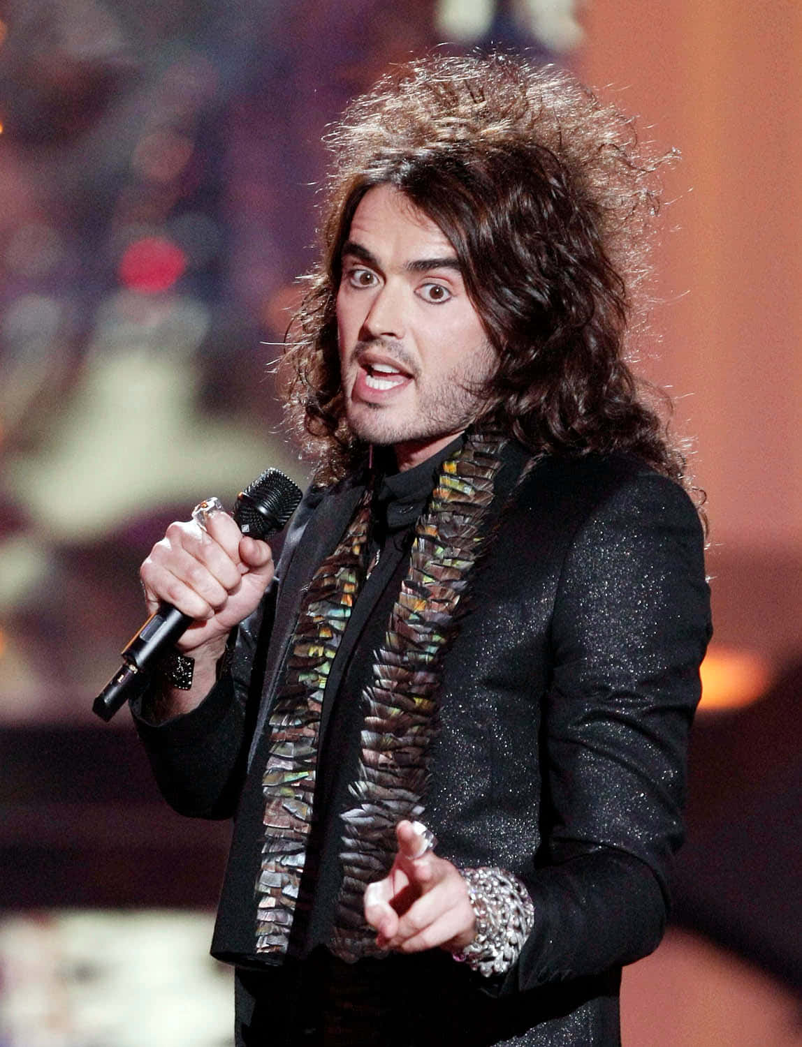 Celebrity 桌布 壁紙 — 一位擁有狂野捲髮的表演者，在燈光明亮的舞台上對著麥克風熱情歌唱。