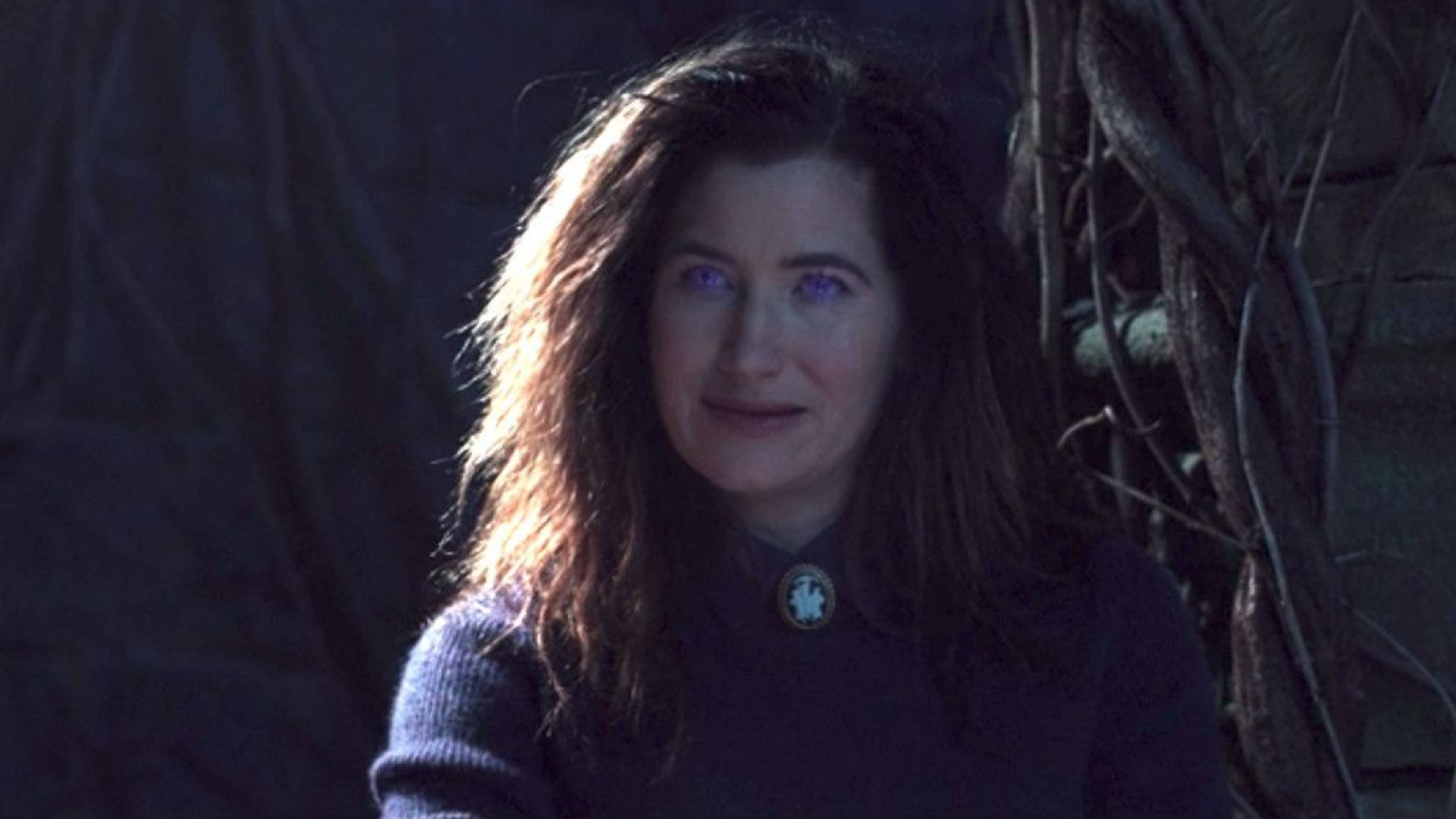 Agatha Harkness 桌布 壁紙 — 一位擁有發光紫色眼睛和深色頭髮的女性站在陰影的樹葉中，穿著帶有圓形胸針的深色毛衣。