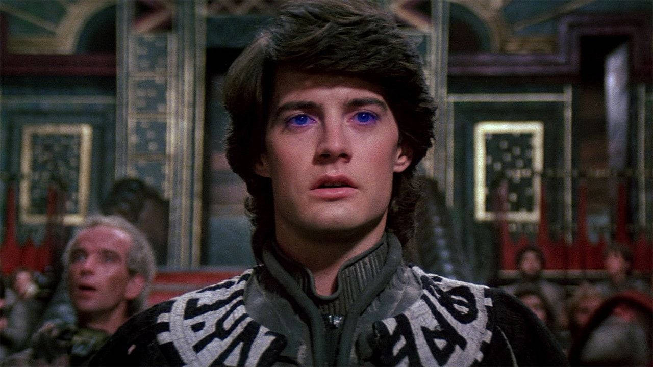Dune Movie 桌布 壁紙 — 一位擁有發光藍色眼睛的年輕男子，在一個裝飾華麗的房間內，背景有其他人，專注地凝視著。