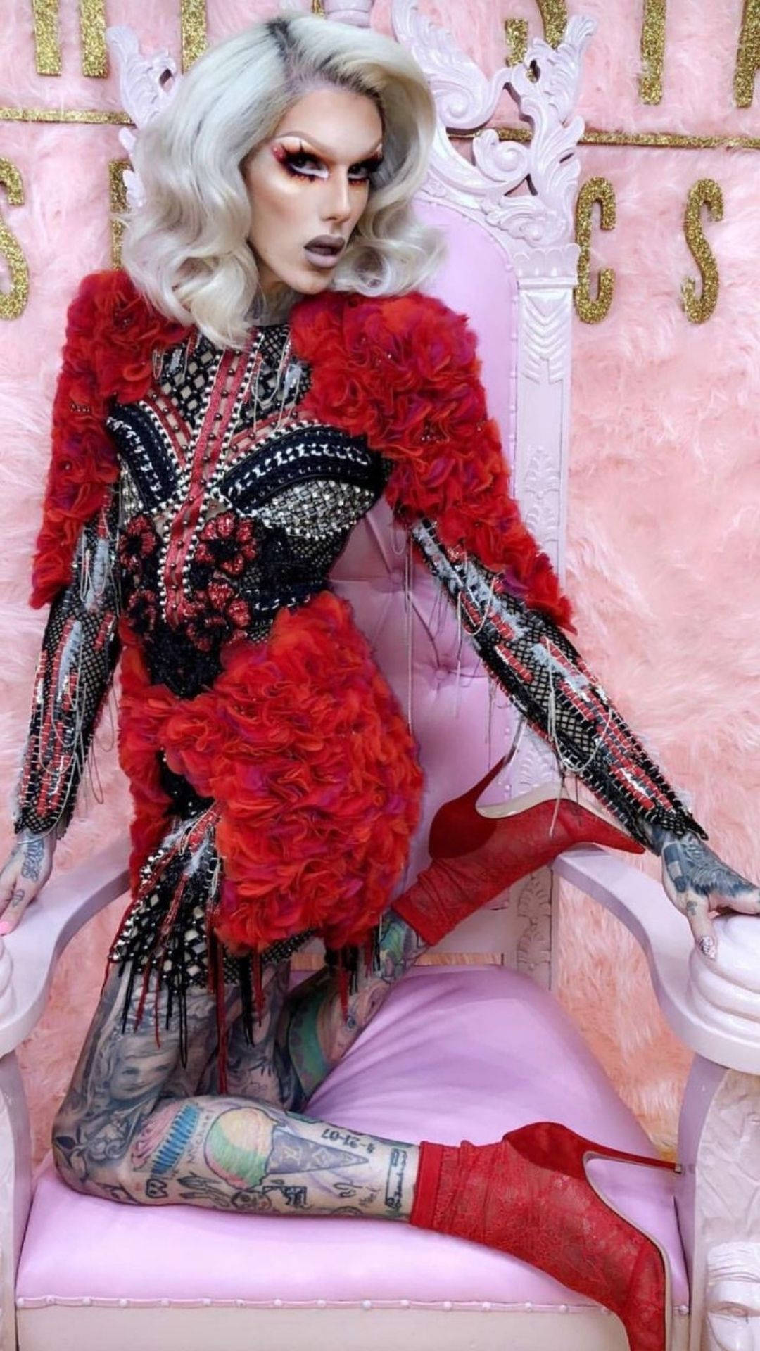 Jeffree Star 桌布 壁紙 — 一位擁有白金髮色和刺青的變裝皇后，身穿紅色羽毛服裝和紅色高跟鞋，威嚴地坐在粉紅色寶座上。