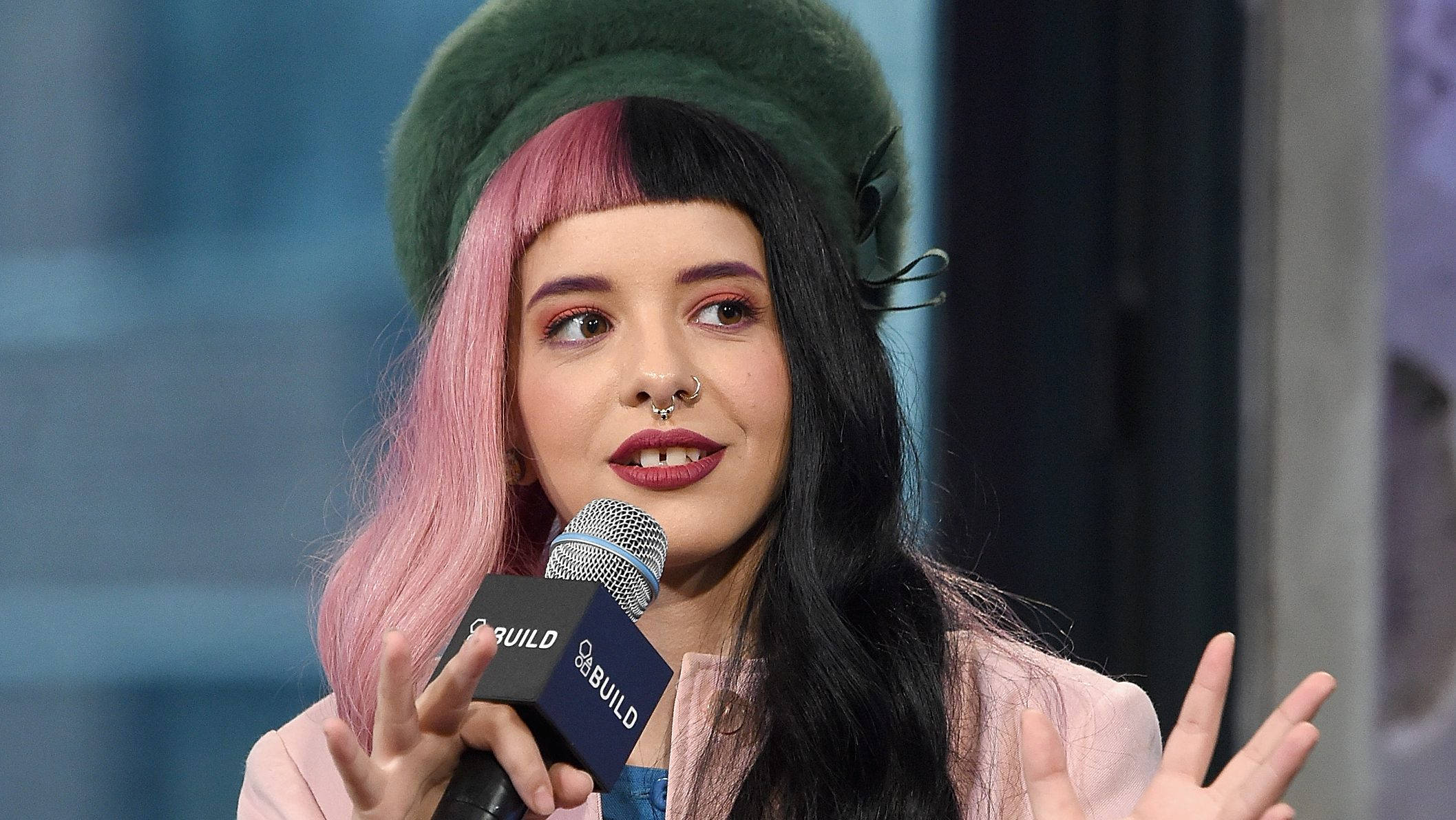 Melanie 桌布 壁紙 — 一位擁有粉紅色和黑色頭髮、戴著綠色貝雷帽的女性，一邊用手勢一邊對著麥克風說話。