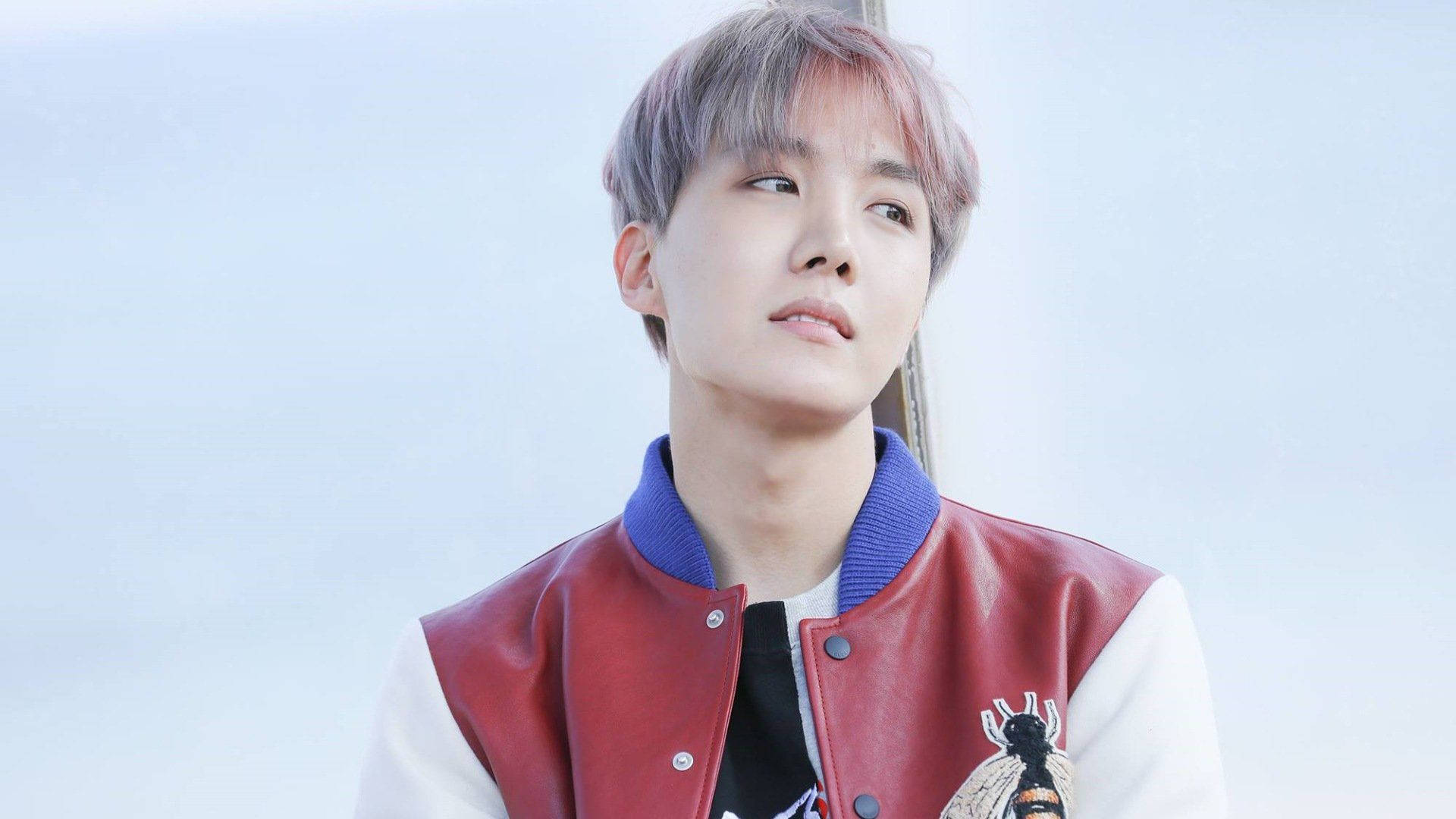 Cute Bts 桌布 壁紙 — 一位擁有粉紅色頭髮的年輕男子，穿著紅白相間的校隊夾克，背景是柔和的藍天。
