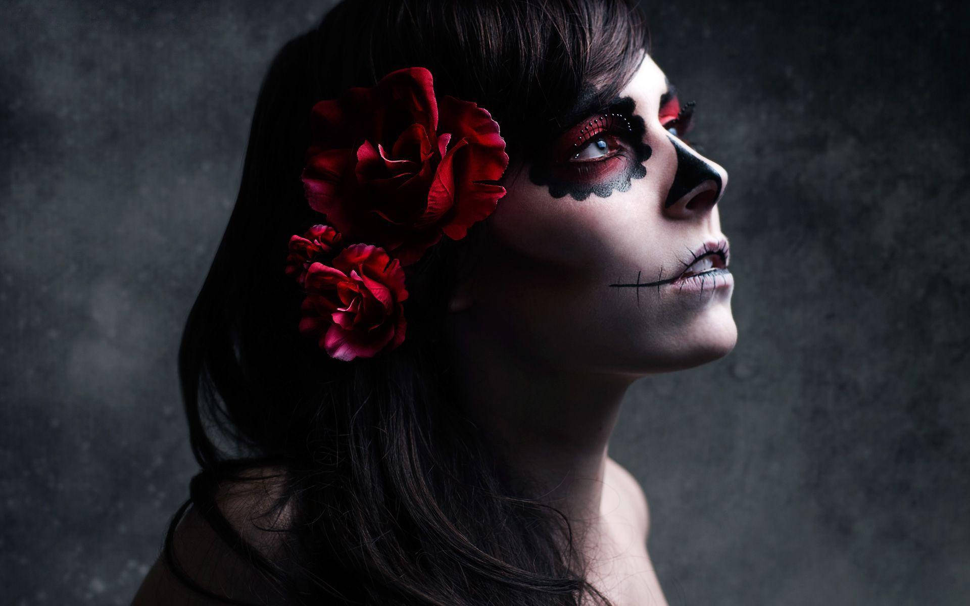Dia De Los Muertos 桌布 壁紙 — 一位擁有精緻糖骷髏妝容、頭髮上插著紅玫瑰的女性，在深色紋理背景前向上凝視。