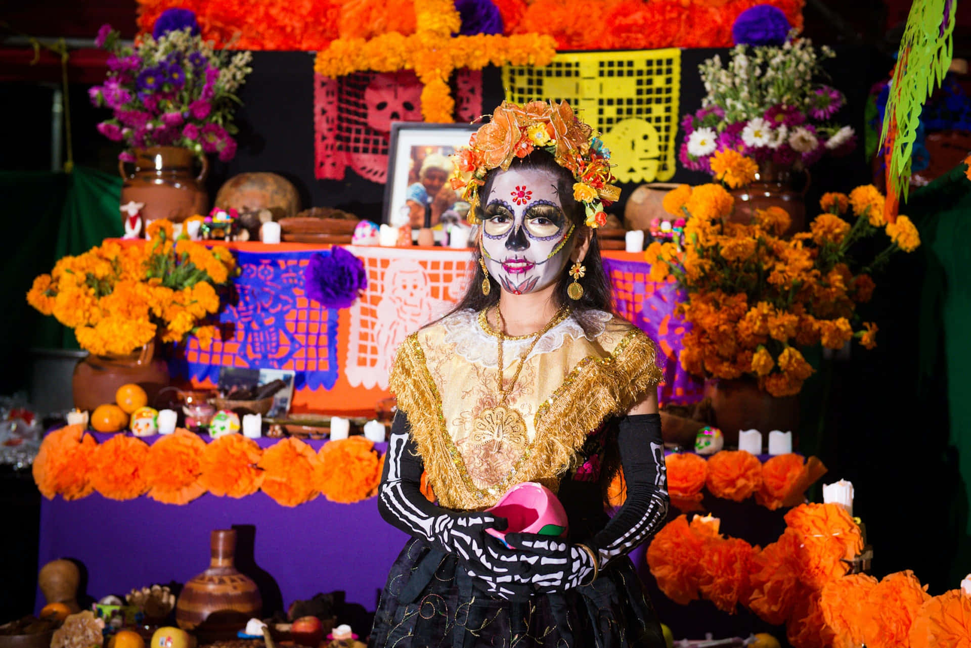 Mexican 桌布 壁紙 — 一位擁有精緻糖骷髏妝容和花冠的女性站在一個充滿活力的祭壇前，祭壇上裝飾著萬壽菊、蠟燭和 papel picado。