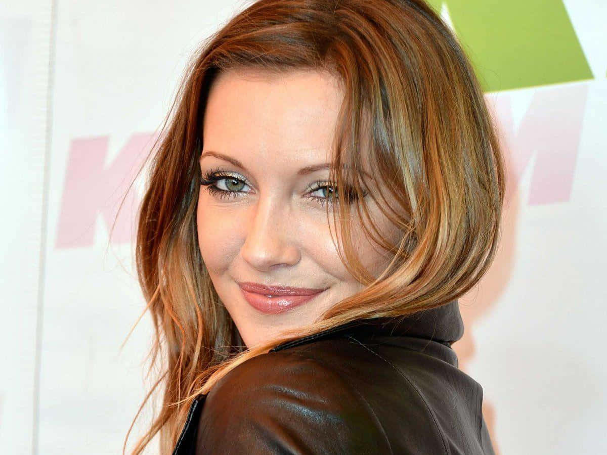 katie cassidy 桌布 壁紙 — 一位擁有紅棕色頭髮和綠色眼睛的女性，穿著棕色皮外套，回頭微笑。