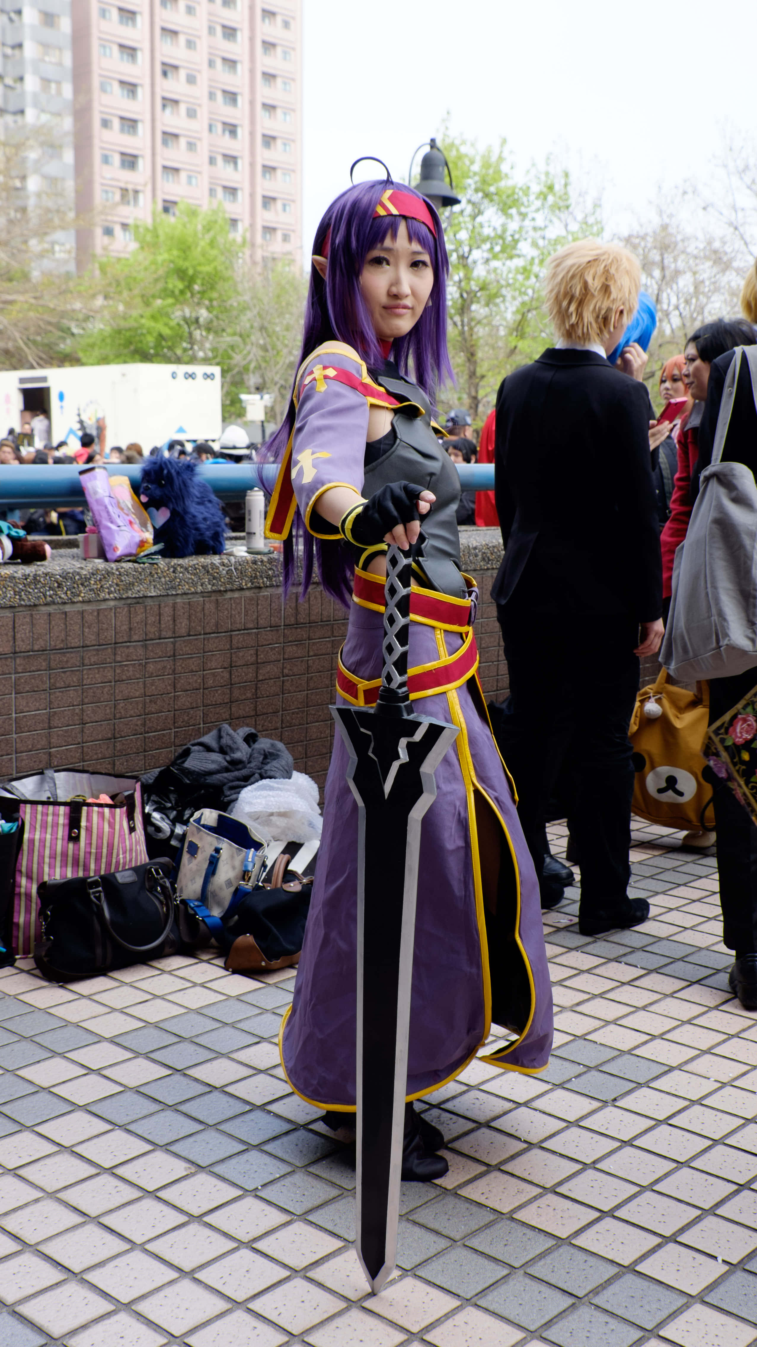 Sword Art Online Cosplay 桌布 壁紙 — 一位擁有紫色頭髮和精靈耳朵的角色扮演者在瓷磚廣場上戶外擺姿勢，手持一把大劍。