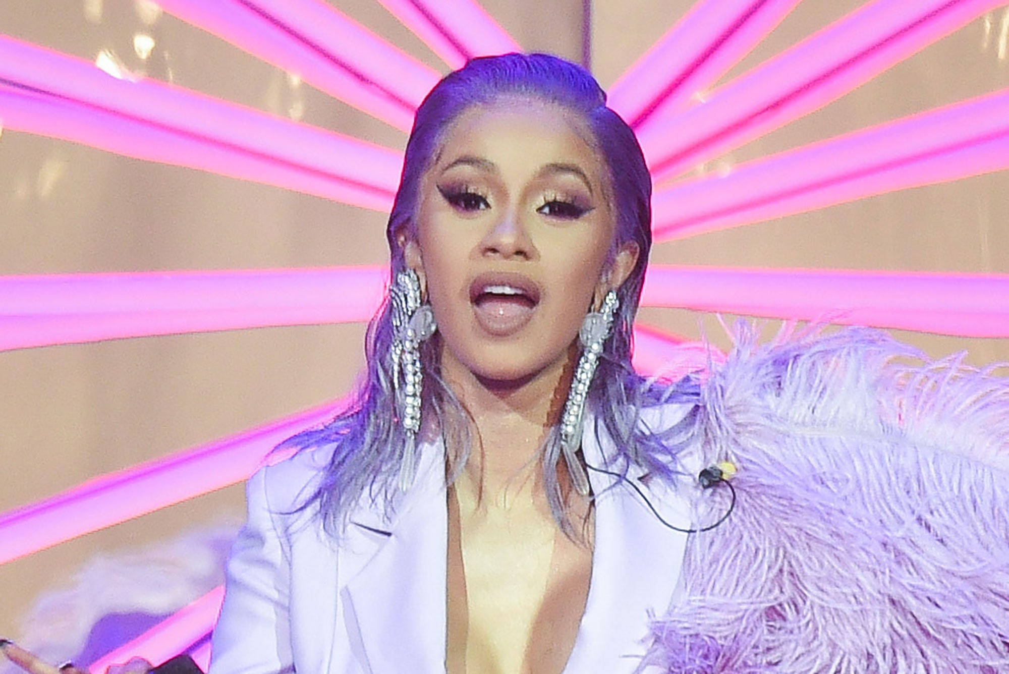 cardi 桌布 壁紙 — 一位擁有紫色頭髮和閃亮耳環的表演者在燦爛的粉紅色燈光背景下熱情歌唱。