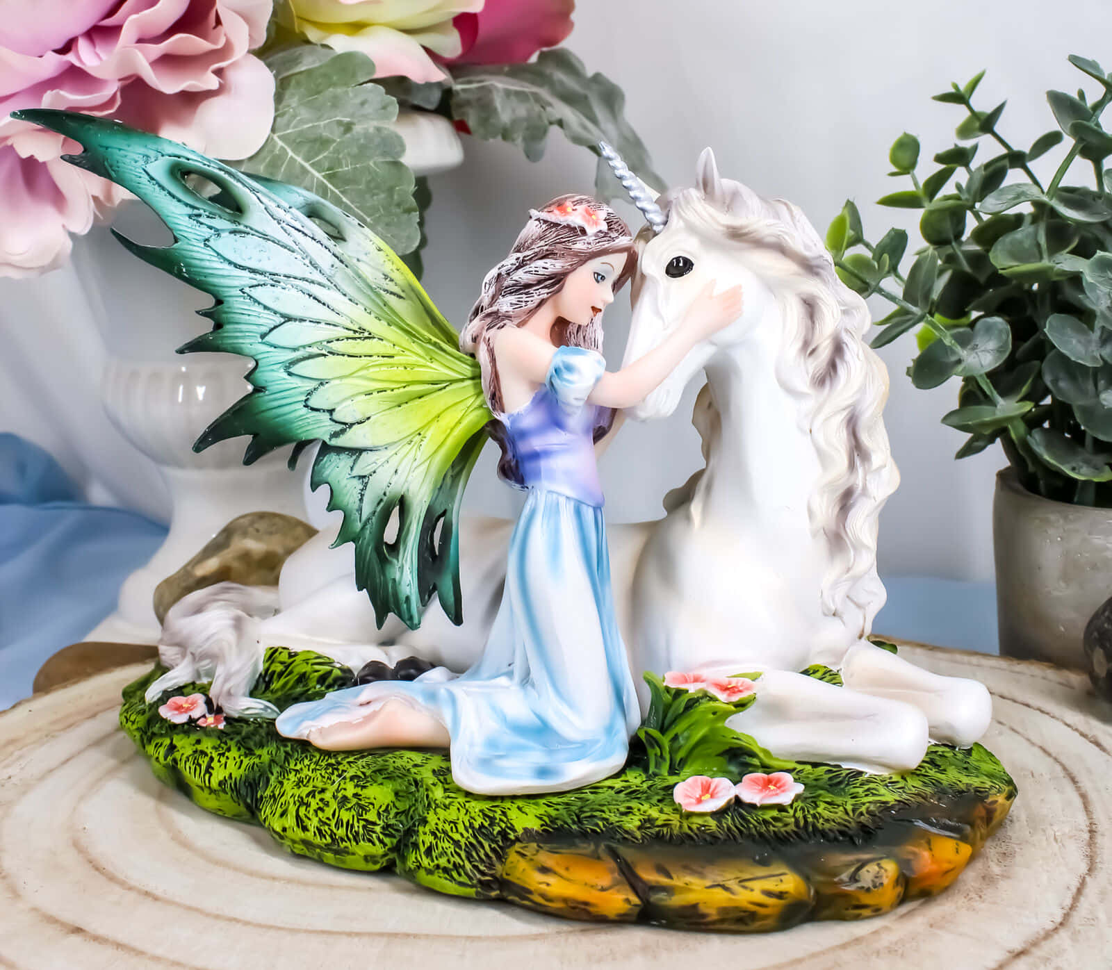 Fairy Wings 桌布 壁紙 — 一位擁有綠色和藍色翅膀的仙女，輕輕撫摸著一隻在長滿粉紅色花朵的草地底座上休息的白色獨角獸。