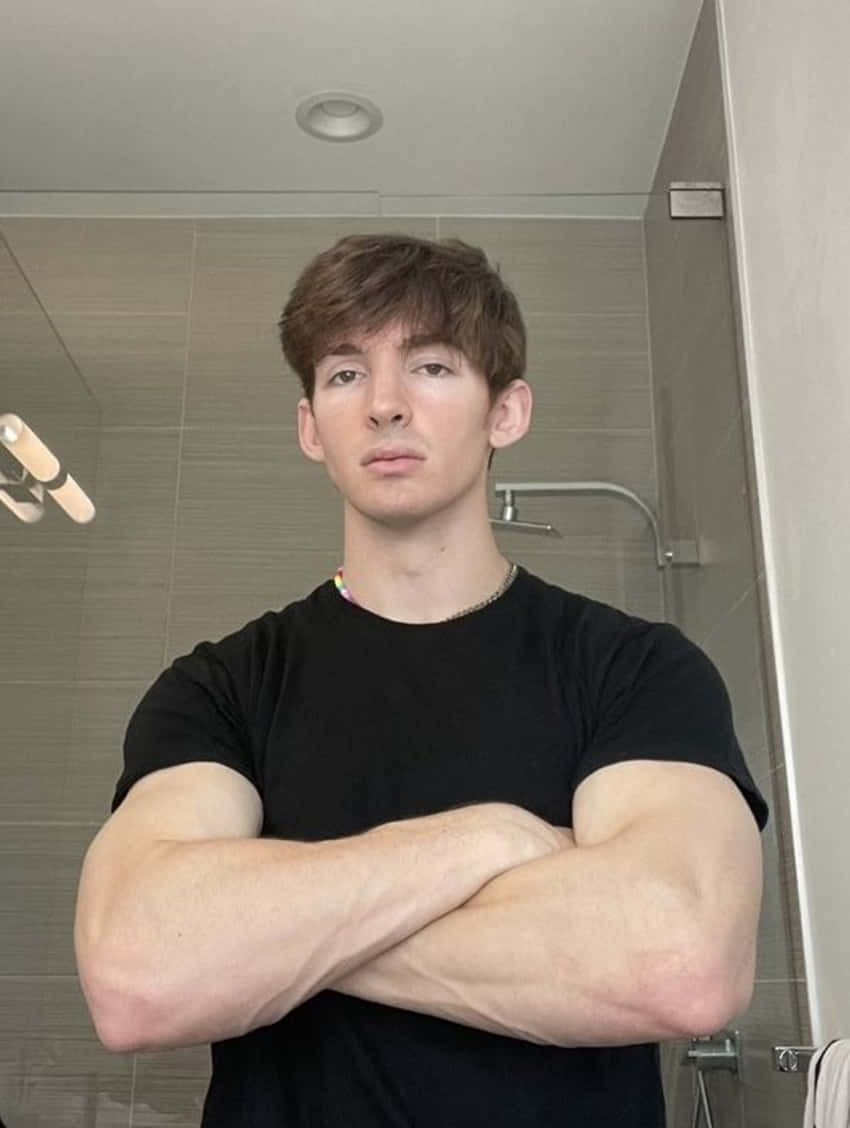 桌布 壁紙 — 一位擁有肌肉手臂的年輕男子自信地站在現代浴室中，雙臂交叉，身穿黑色T恤。