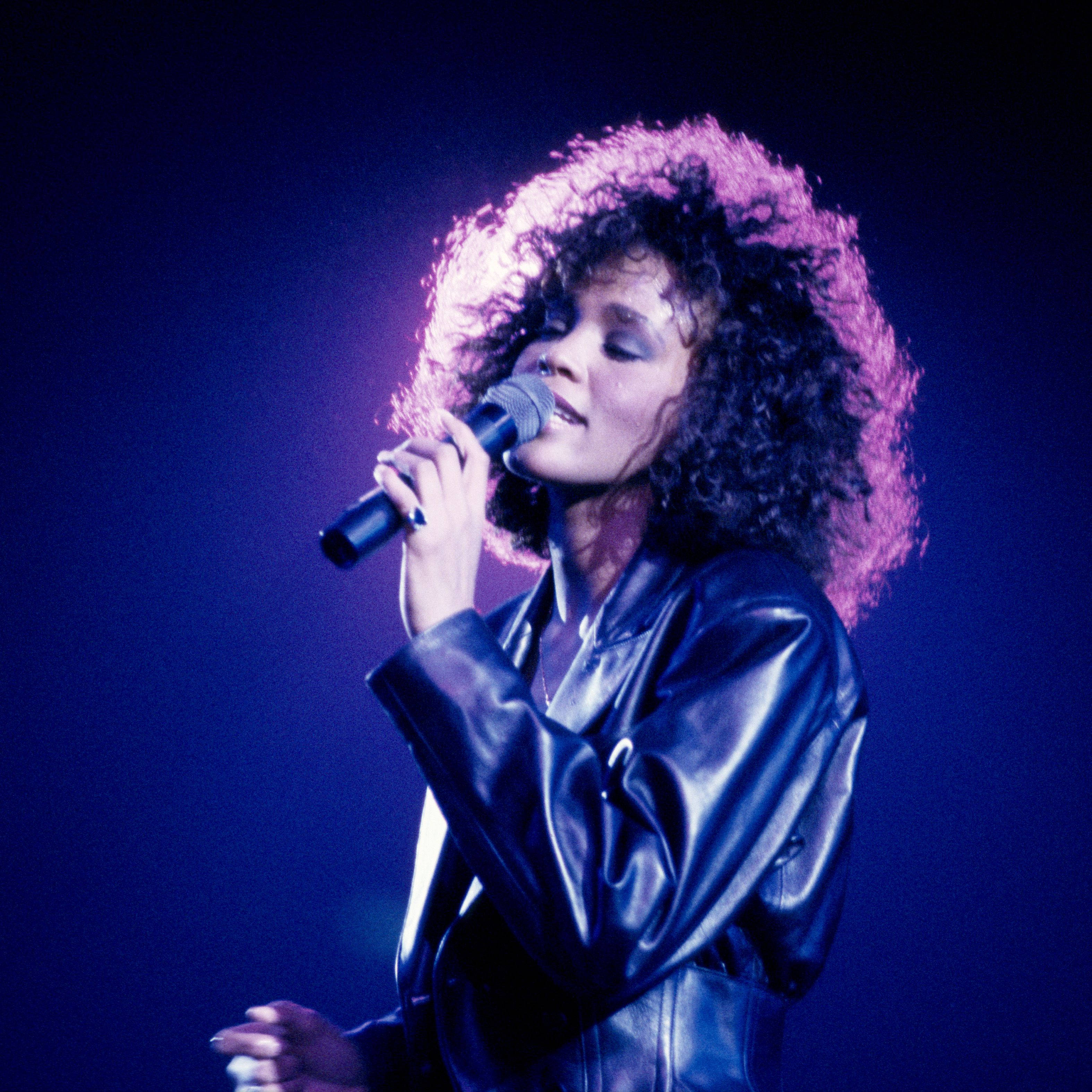 Whitney 桌布 壁紙 — 一位擁有蓬鬆捲髮的歌手沐浴在紫色燈光下，穿著閃亮的黑色皮夾克，對著麥克風熱情演唱。