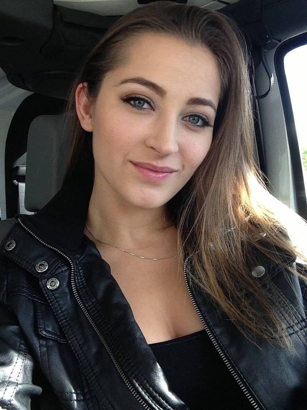 Dani Daniels 桌布 壁紙 — 一位擁有藍色眼睛和長棕色頭髮的年輕女性，在車內穿著黑色皮夾克，溫柔地微笑。