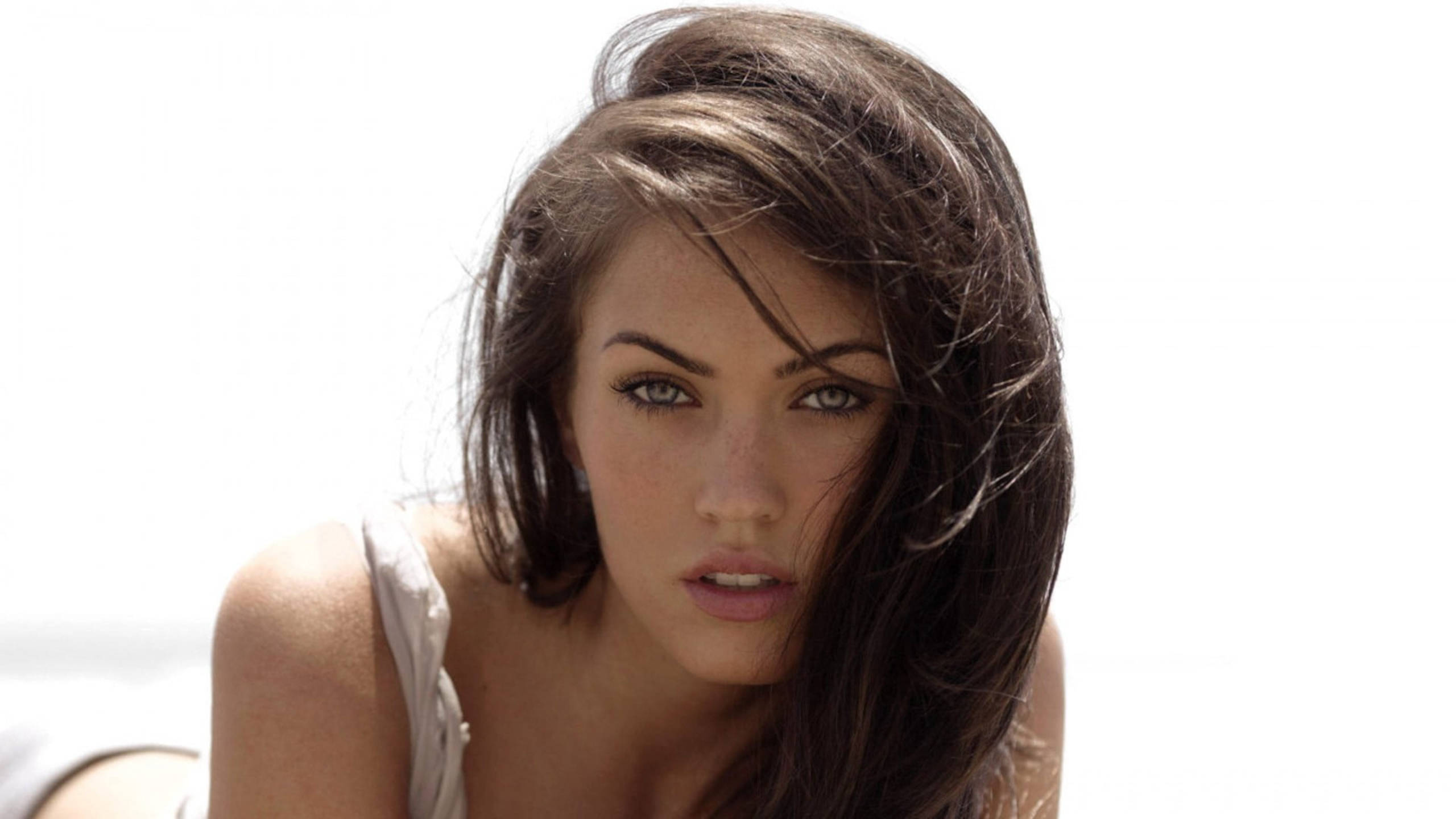 Megan Fox 桌布 壁紙 — 一位擁有被風吹亂深色頭髮和強烈眼神的女性，在明亮的白色背景下直視鏡頭。