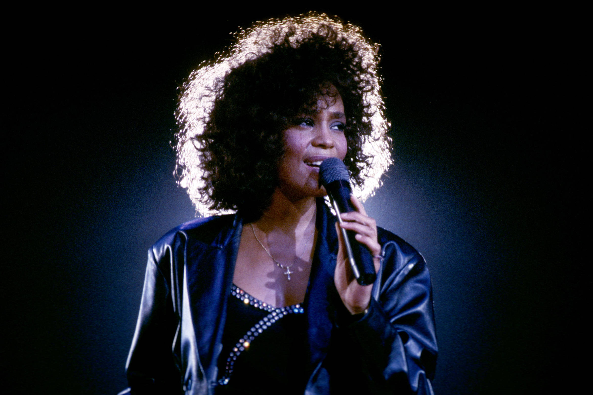 Whitney 桌布 壁紙 — 一位擁有豐滿非洲髮型的女性在黑暗背景下，於戲劇性的舞台燈光下對著麥克風唱歌。