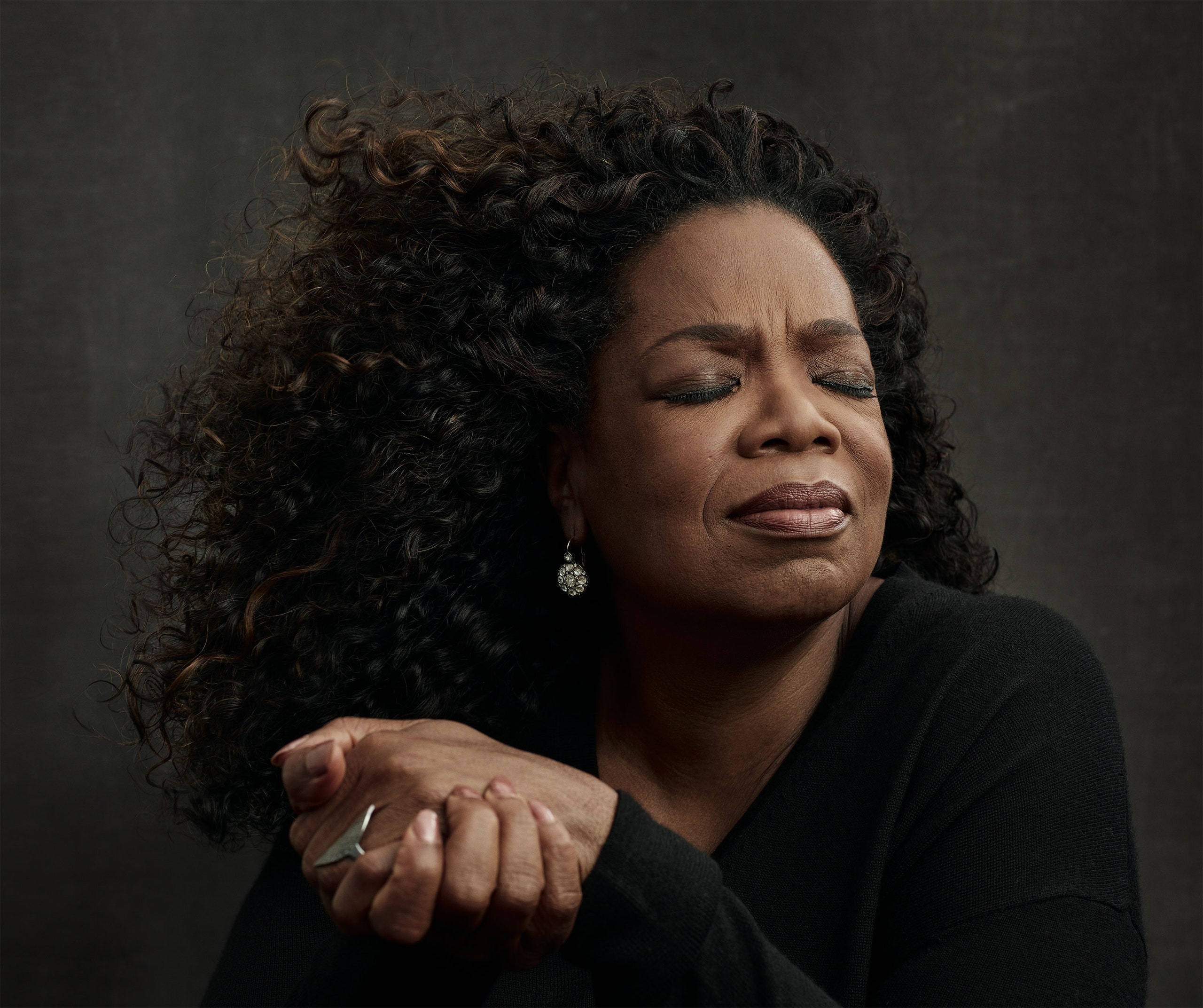 Oprah 桌布 壁紙 — 一位擁有豐盈捲髮、閉著眼睛的女性，將交疊的雙手放在胸前，背景為深色紋理。