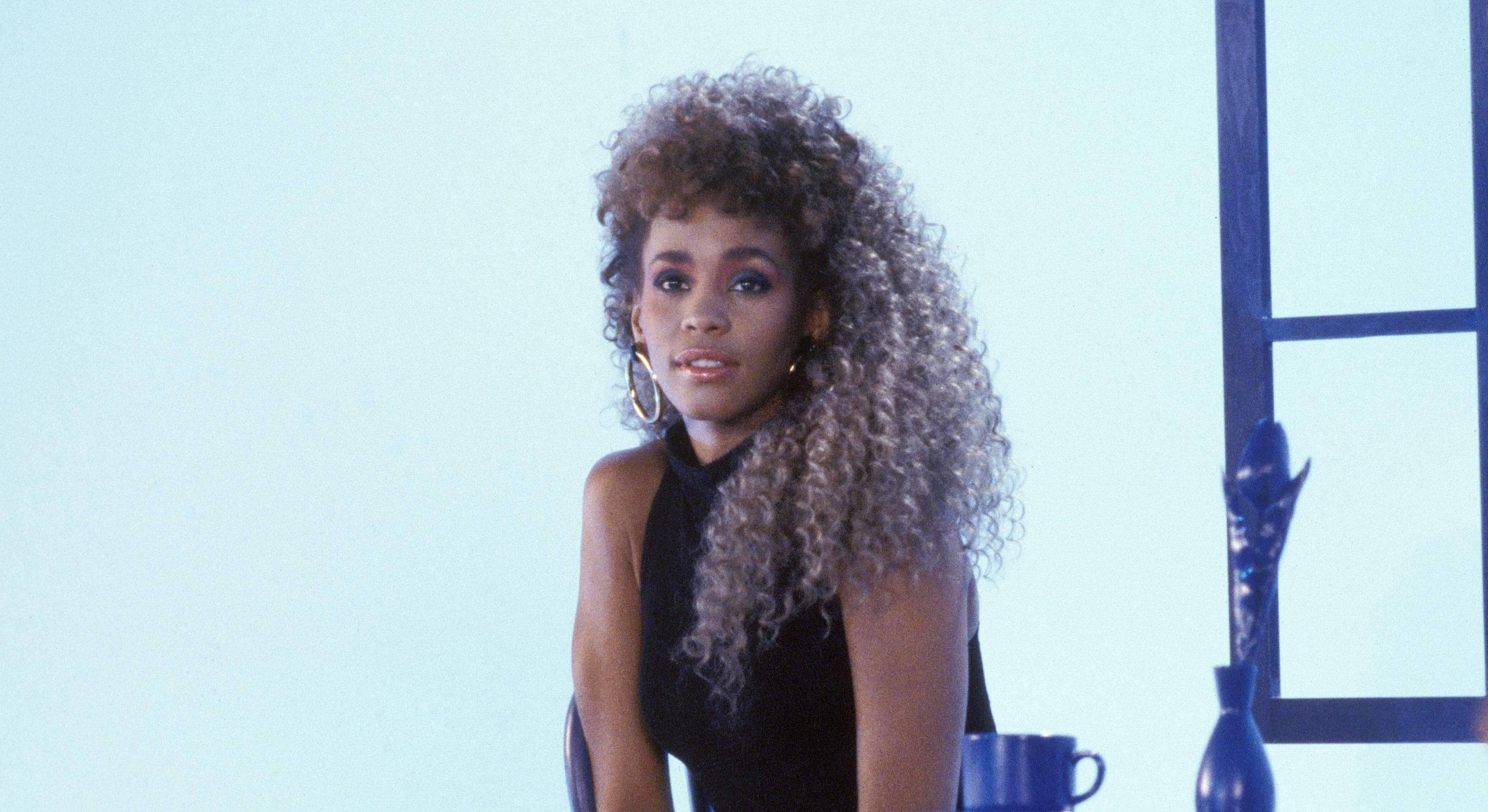 Whitney 桌布 壁紙 — 一位擁有豐盈捲髮和圈狀耳環的女性自信地坐在帶有藍色點綴的簡約房間中。
