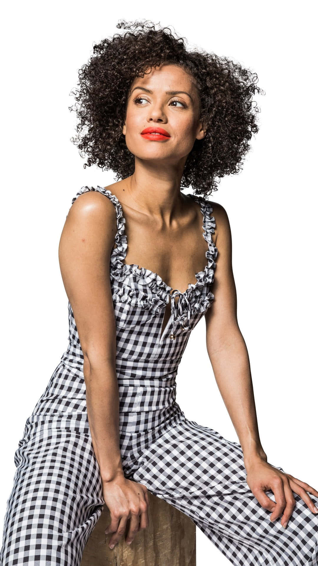 Gugu Mbatha-raw 桌布 壁紙 — 一位擁有豐盈捲髮和大膽紅唇的女性，自信地穿著黑白格紋連身褲，坐在純白背景前。