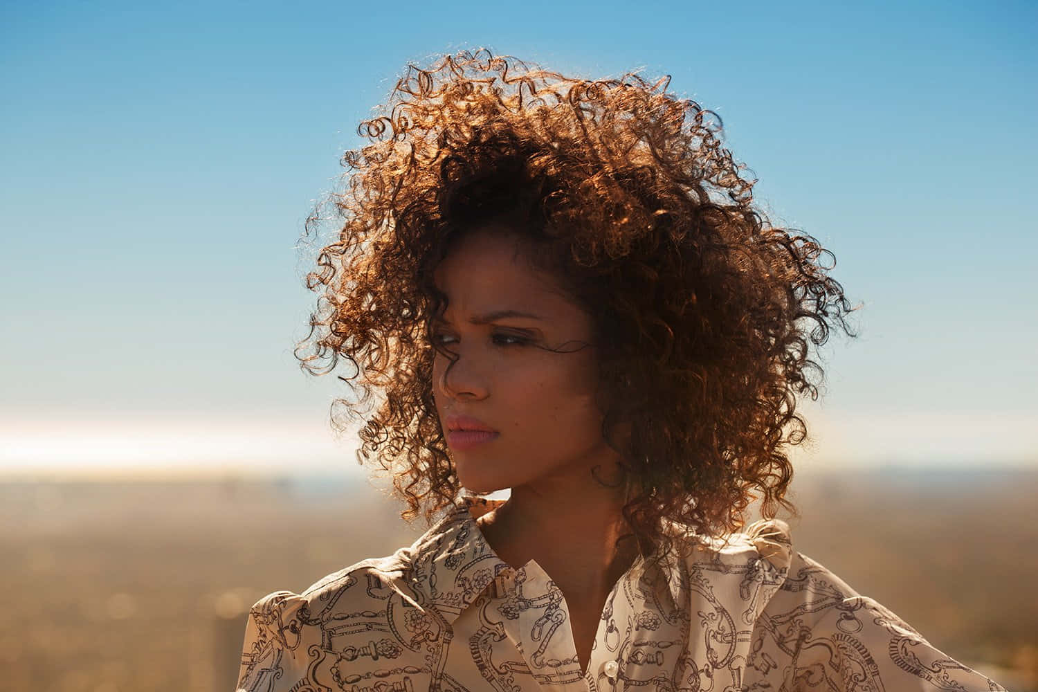 Gugu Mbatha-raw 桌布 壁紙 — 一位擁有豐盈捲髮的女性，在朦朧的遠景之上，對著清澈的藍天若有所思地凝視。