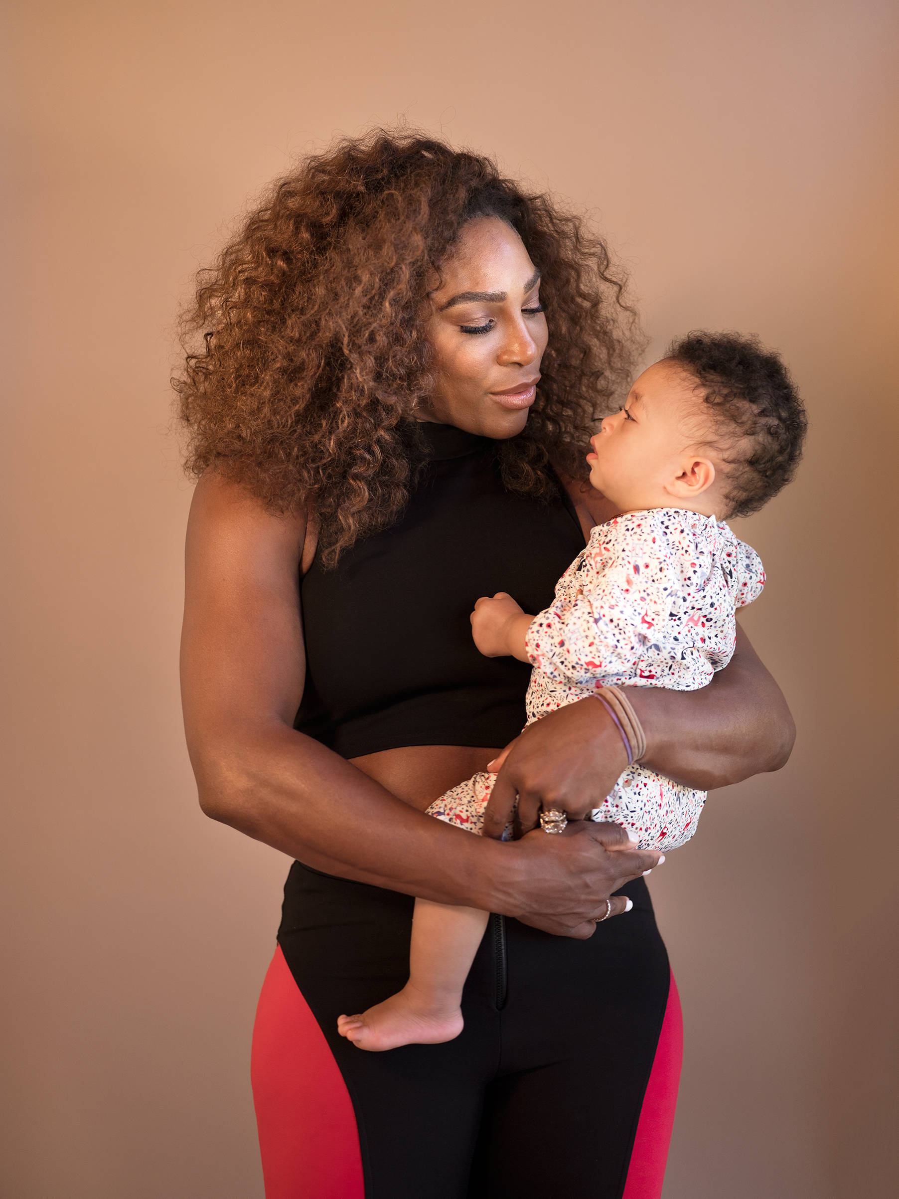 Serena 桌布 壁紙 — 一位擁有豐盈捲髮的女性，在溫暖的米色背景下，懷抱著穿著花卉連體衣的嬰兒。