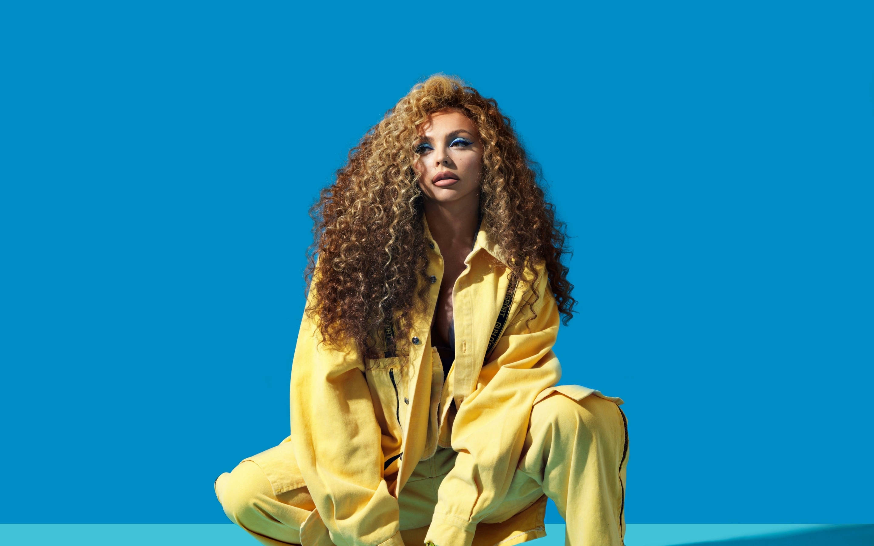 Jesy Nelson 桌布 壁紙 — 一位擁有豐盈捲髮的女性，身穿亮黃色服裝，坐在鮮豔的藍色背景前。