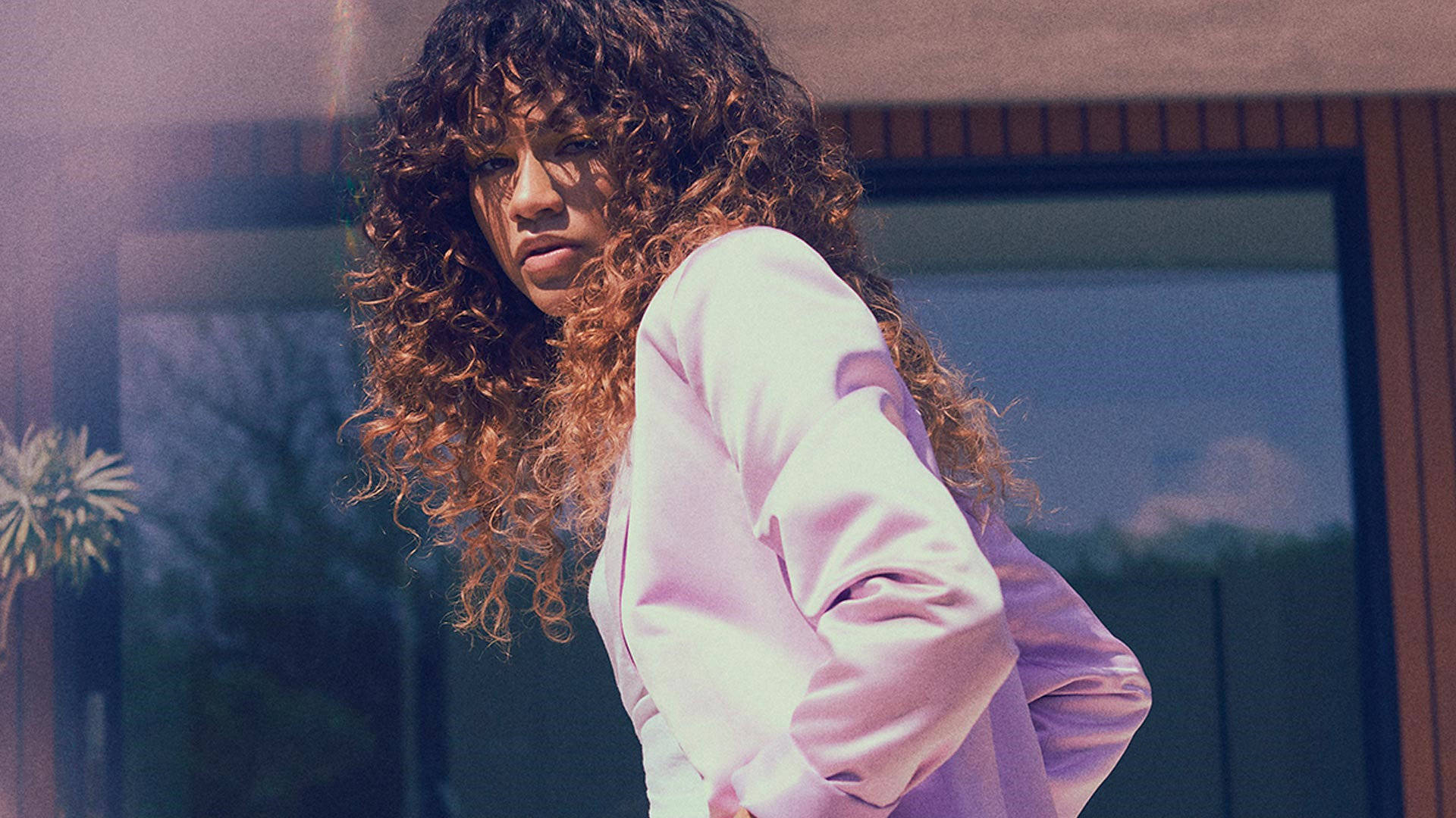 Zendaya 桌布 壁紙 — 一位擁有豐盈捲髮的女性穿著柔軟的粉紅色襯衫，站在現代玻璃建築附近的戶外。