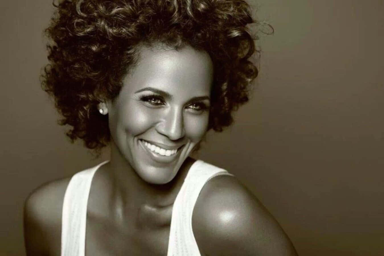 Nicole Ari Parker 桌布 壁紙 — 一位擁有豐盈捲髮的容光煥發女性，在暖色調的攝影棚肖像中穿著白色背心，閃耀光芒。