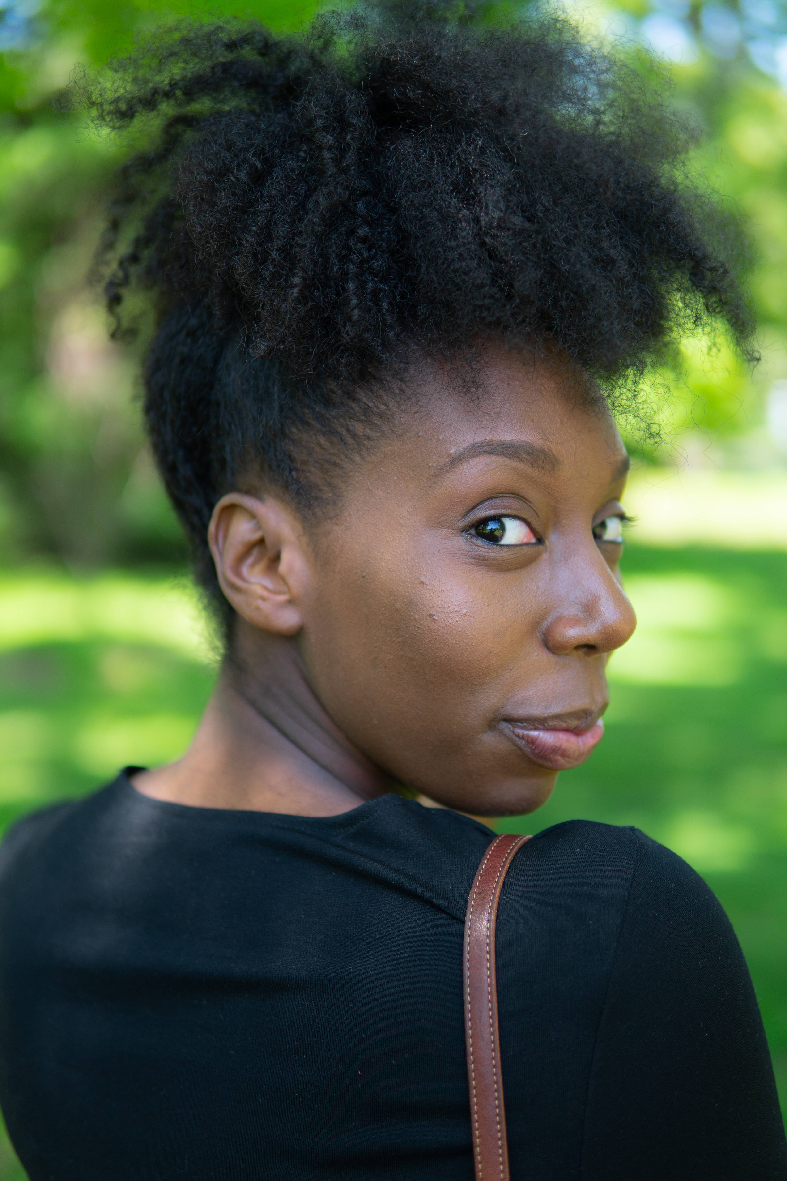 Black Girl 桌布 壁紙 — 一位擁有豐盈捲髮的年輕女性，在明亮綠色模糊的公園背景下，轉頭越過肩膀。