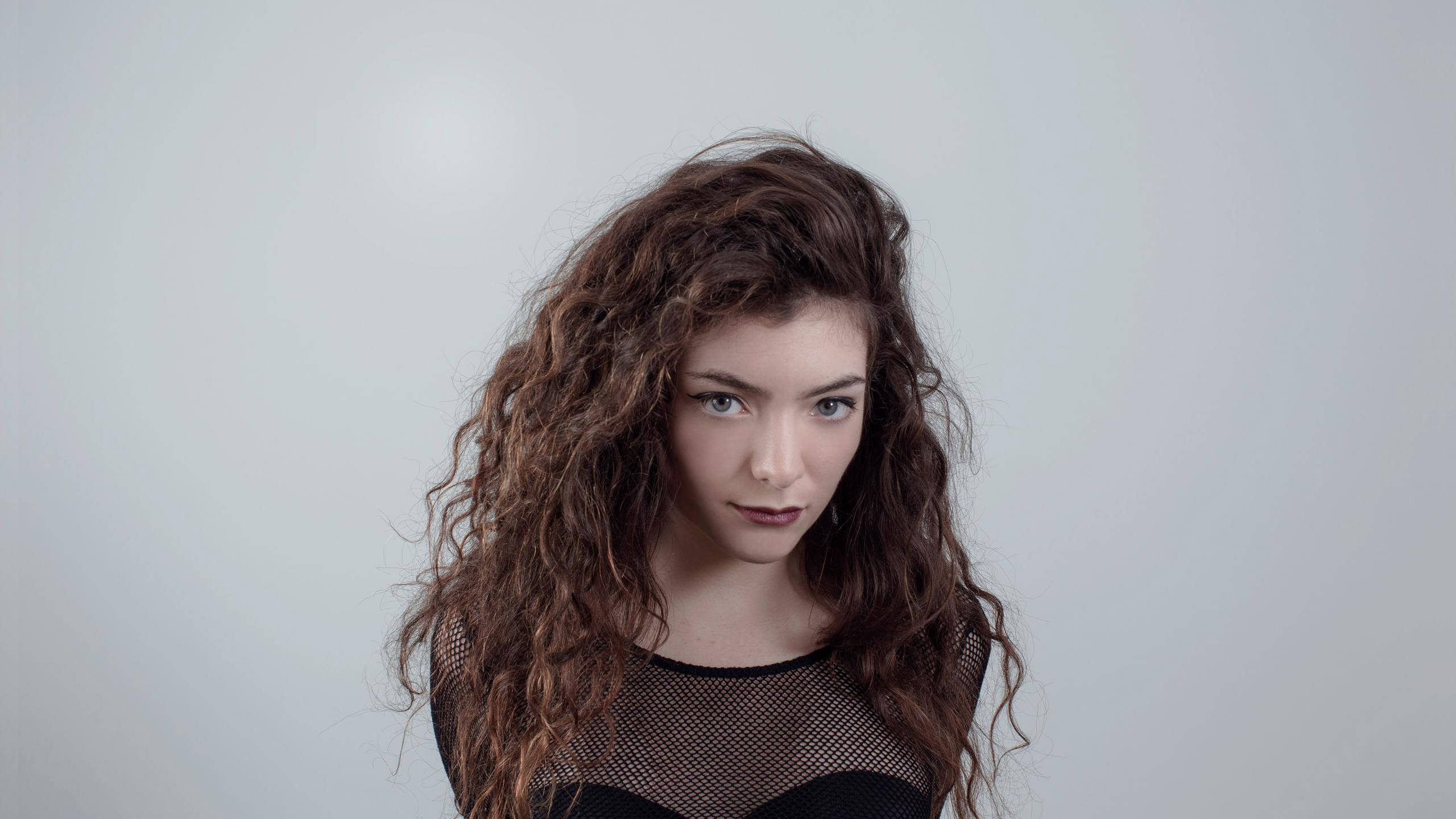 Lorde 桌布 壁紙 — 一位擁有豐盈棕色捲髮和引人注目的藍眼睛的女性，在淺灰色純色背景下直視鏡頭。