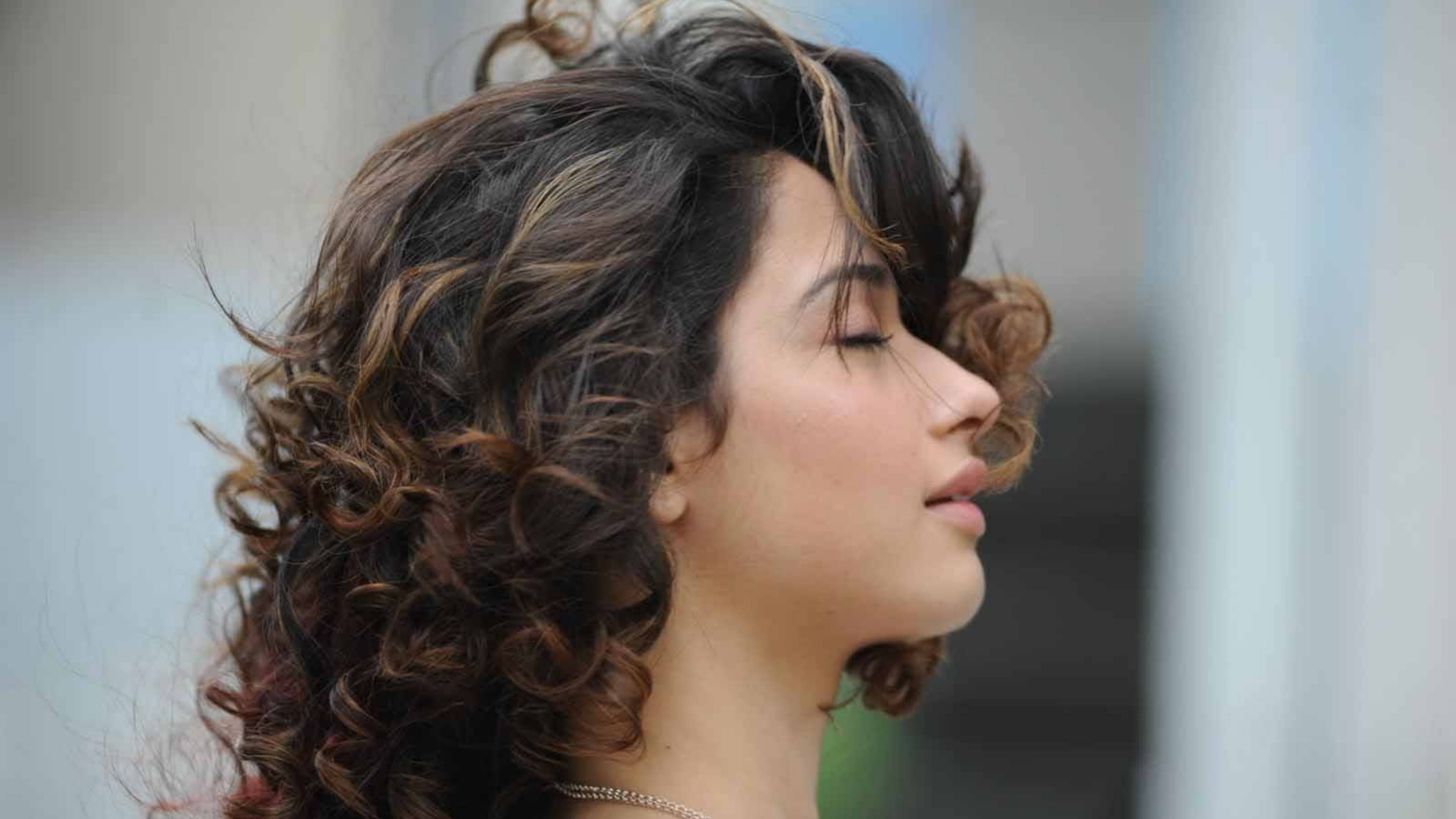 tamannaah bhatia 桌布 壁紙 — 一位擁有豐盈棕色捲髮的女性，在柔和模糊的城市背景下閉上眼睛，安然休憩。