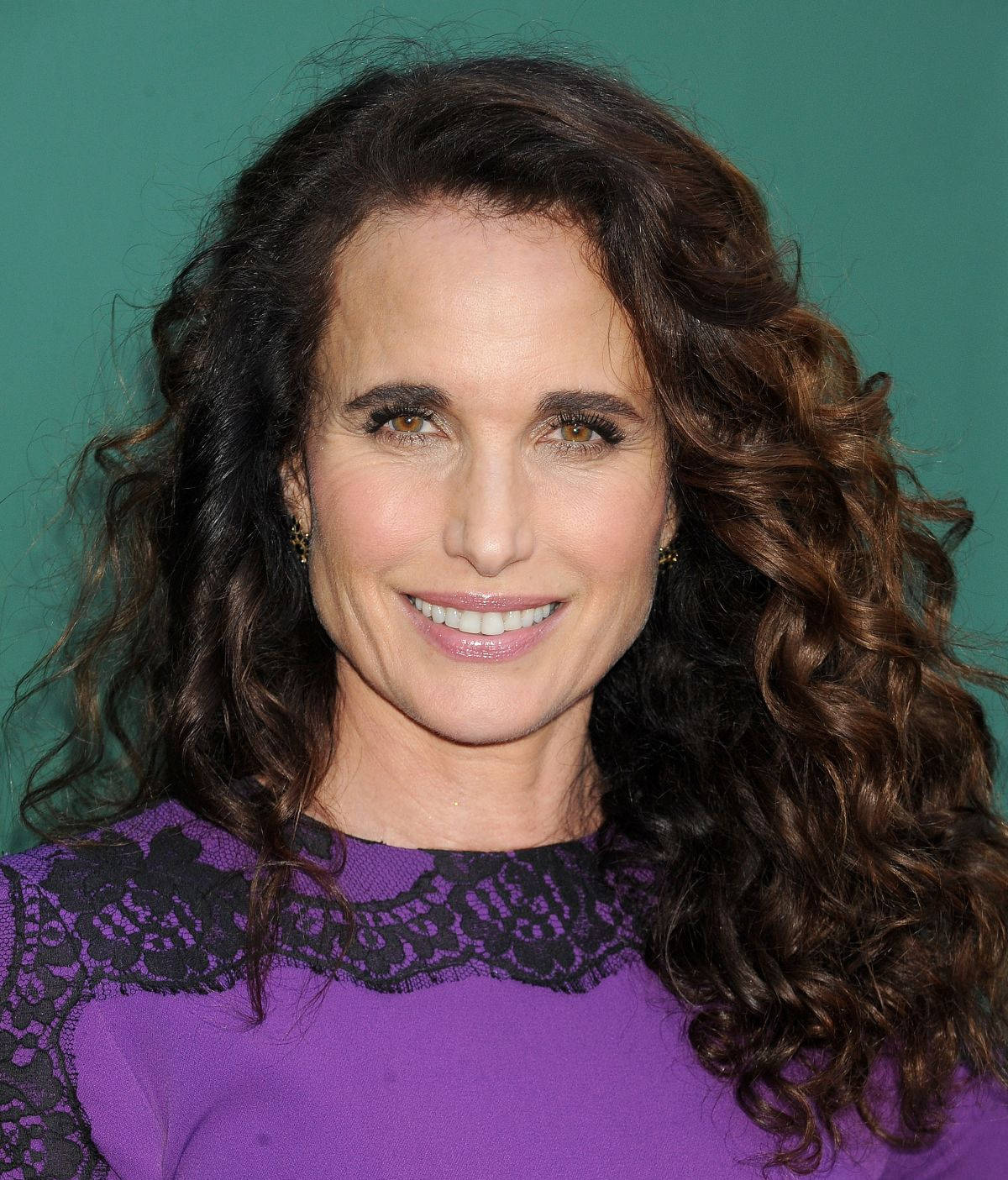 andie macdowell 桌布 壁紙 — 一位擁有豐盈棕色捲髮的女性，在純青色背景前溫暖微笑，穿著紫色蕾絲邊上衣。