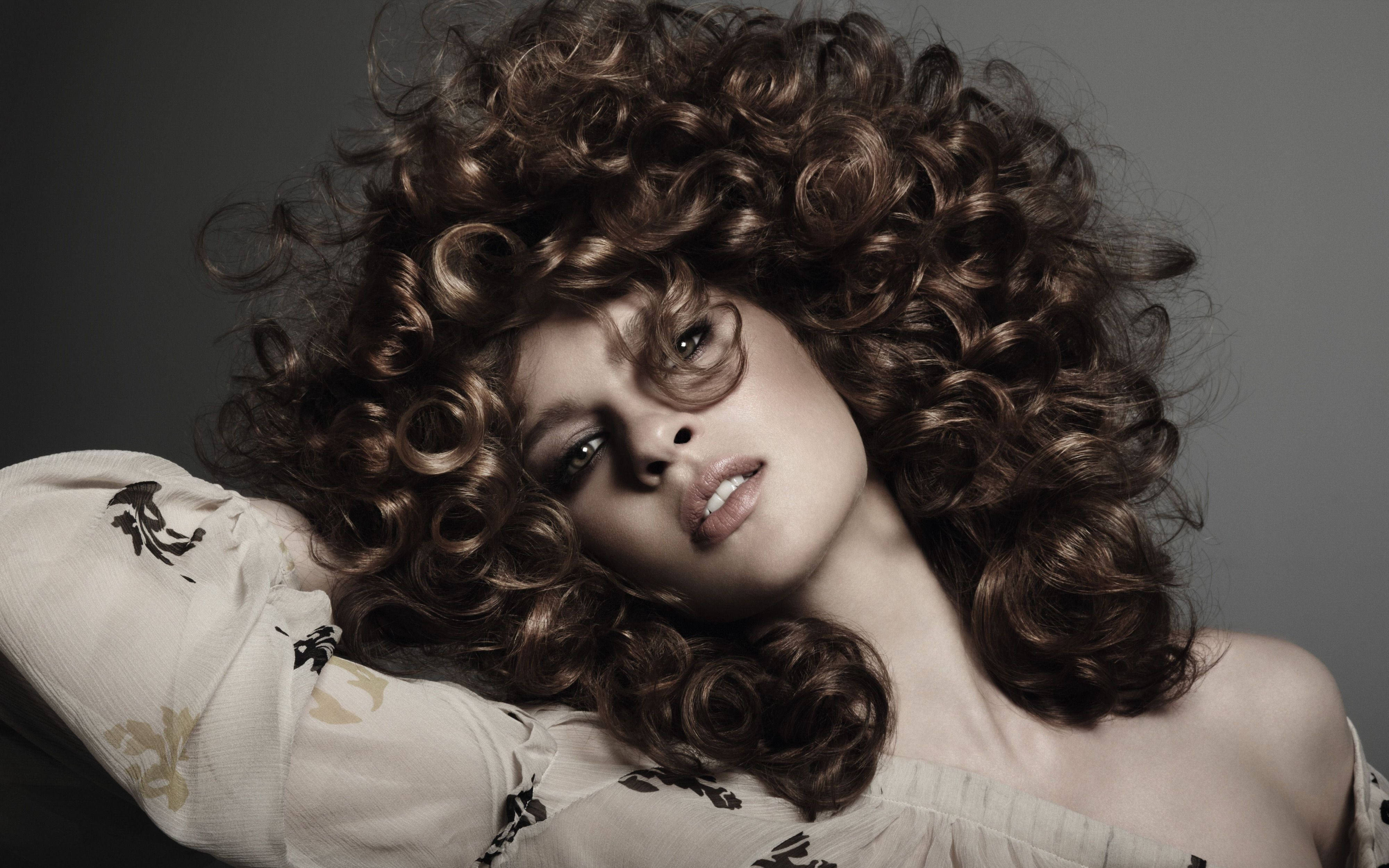 Curly Hair 桌布 壁紙 — 一位擁有豐盈棕色捲髮的女性向後靠著灰色背景，眼神強烈，嘴唇微張。
