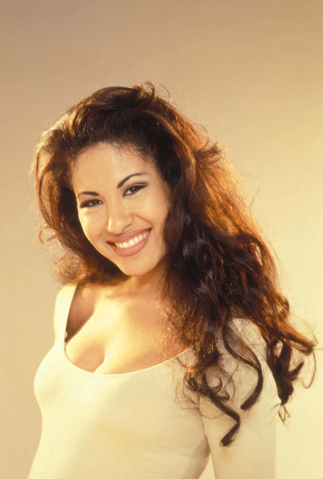 Selena Quintanilla iPhone 桌布 壁紙 — 一位擁有豐盈棕色捲髮的容光煥發的女性，在柔和的米色工作室背景下溫暖地微笑。