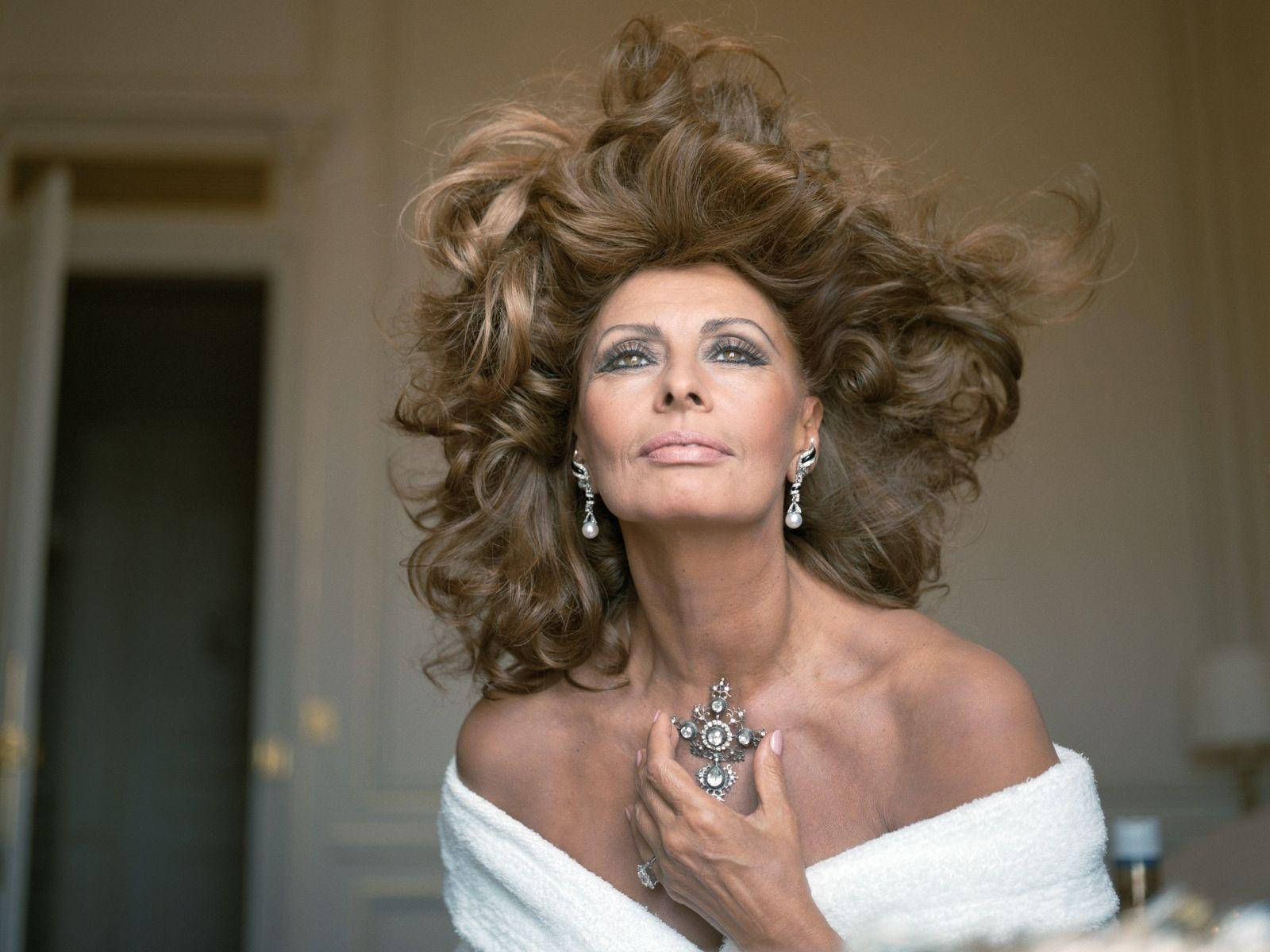 Sophia Loren 桌布 壁紙 — 一位擁有豐盈棕色頭髮和鑽石珠寶的迷人女性，在室內自信地穿著白色長袍擺姿勢。