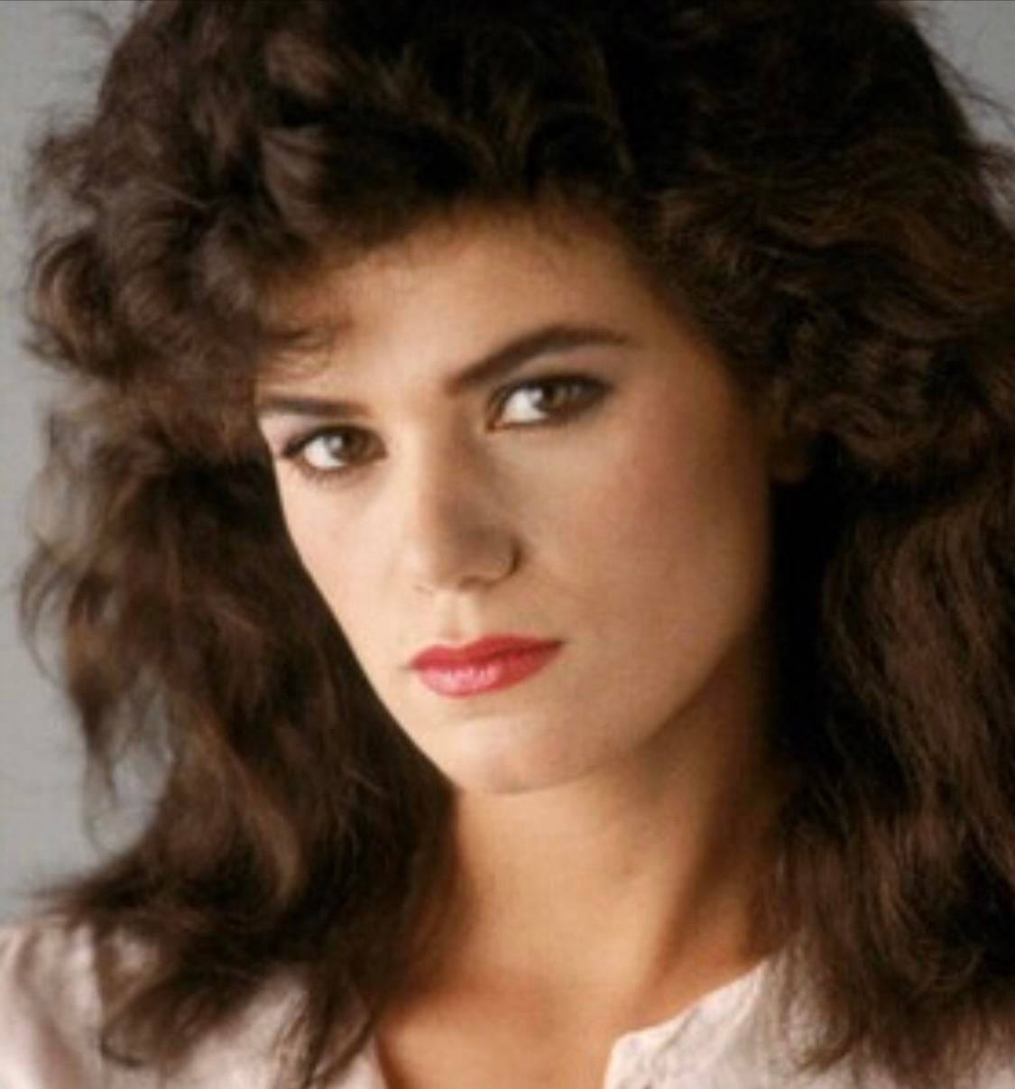Actress HD 桌布 壁紙 — 一位擁有豐盈深色捲髮和大膽紅唇的女性，在灰色中性背景下強烈凝視。